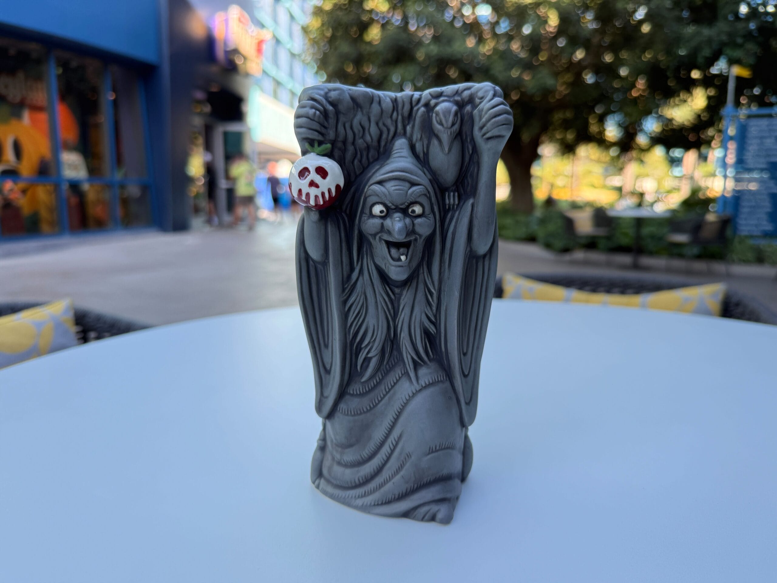 A gray witch figurine holding an Evil Queen Mug stands on a white table outdoors, with trees and buildings behind.