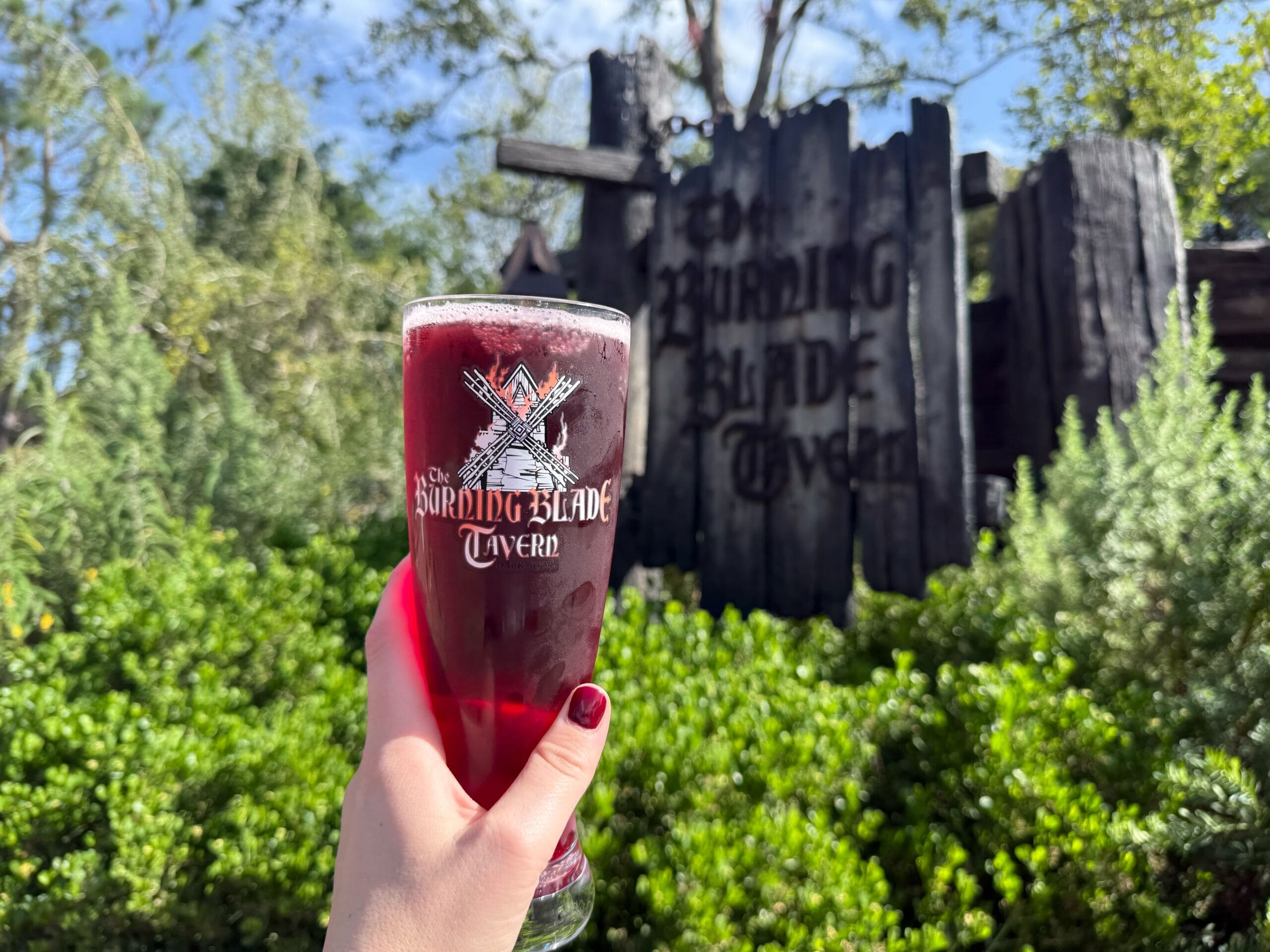 A hand holds a glass of dark red drink in front of a rustic "Burning Blade Tavern" sign surrounded by greenery.