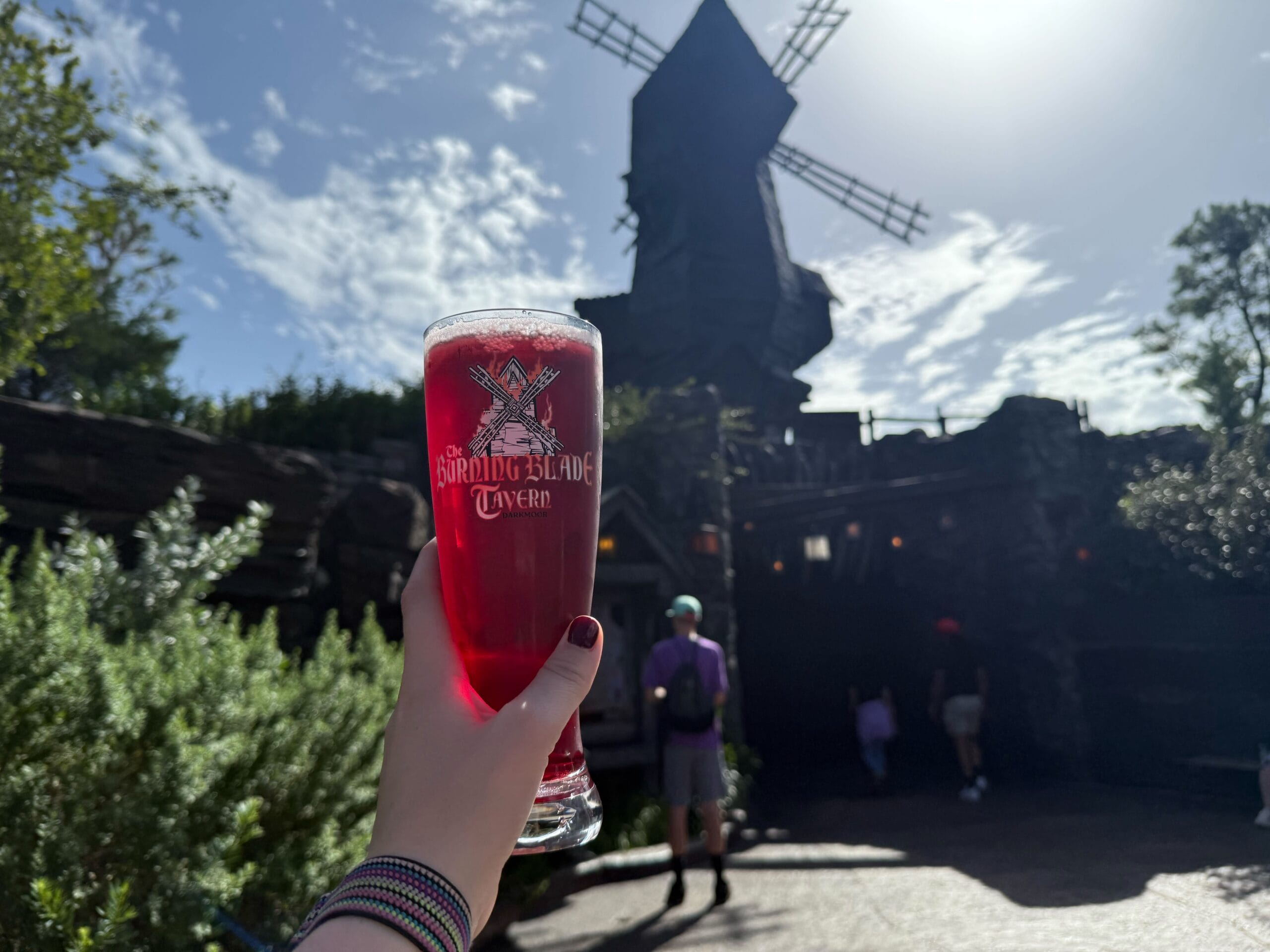 A hand holds a tall glass of red drink in front of a windmill building under a partly cloudy sky.