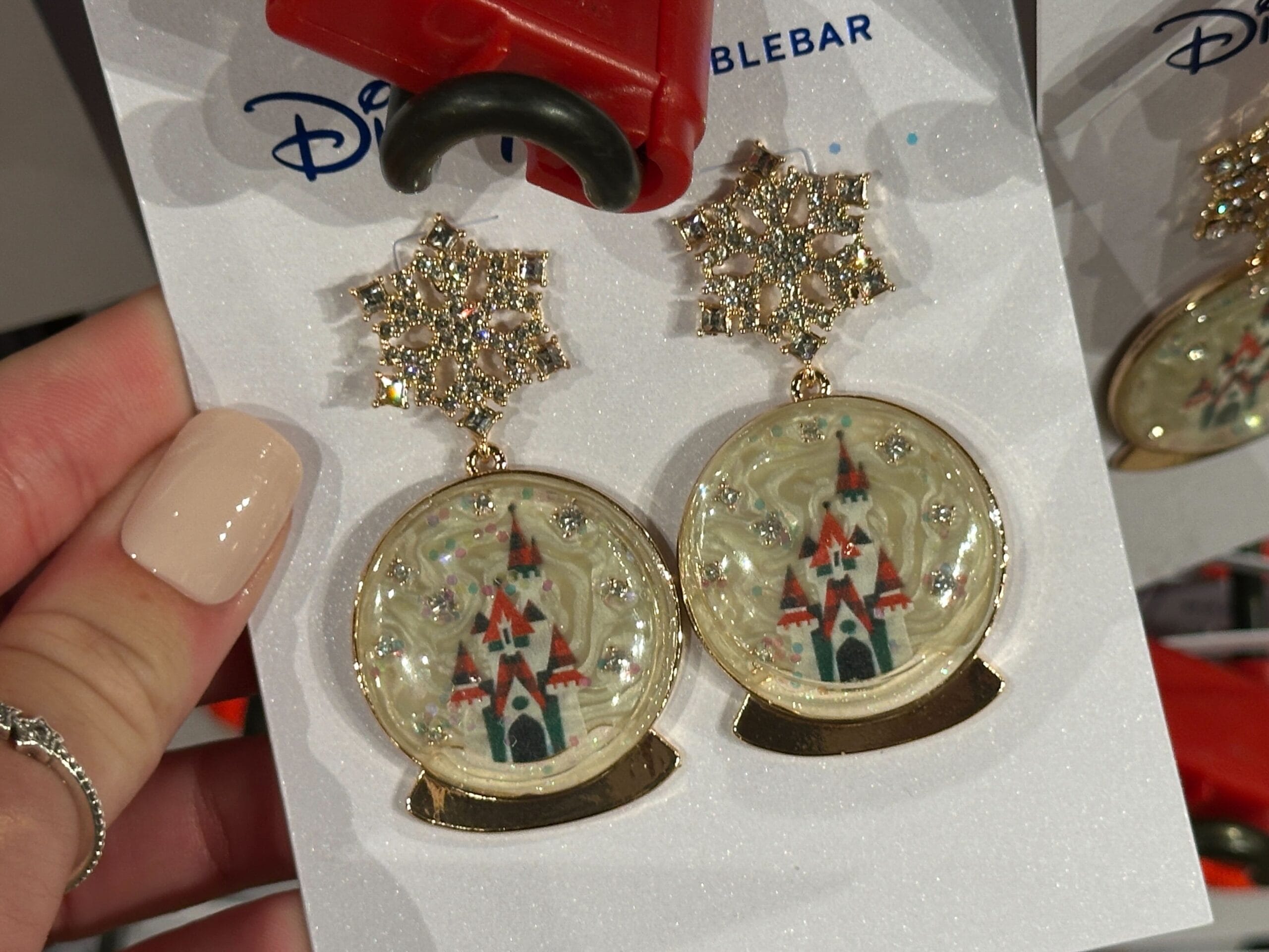 Hand holding earrings shaped like snow globes featuring Cinderella Castle, distinctive of Walt Disney World, with gold snowflake tops.