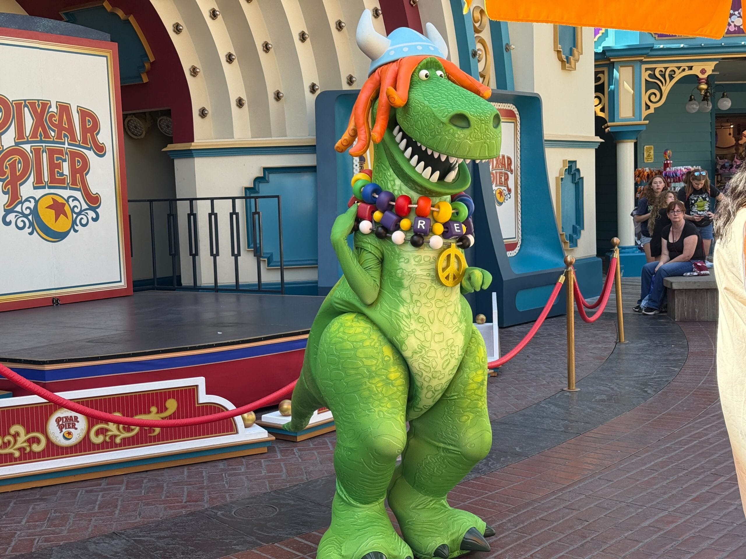 Rex the dinosaur in a colorful hippie costume stands near a stage at Pixar Pier in a theme park.
