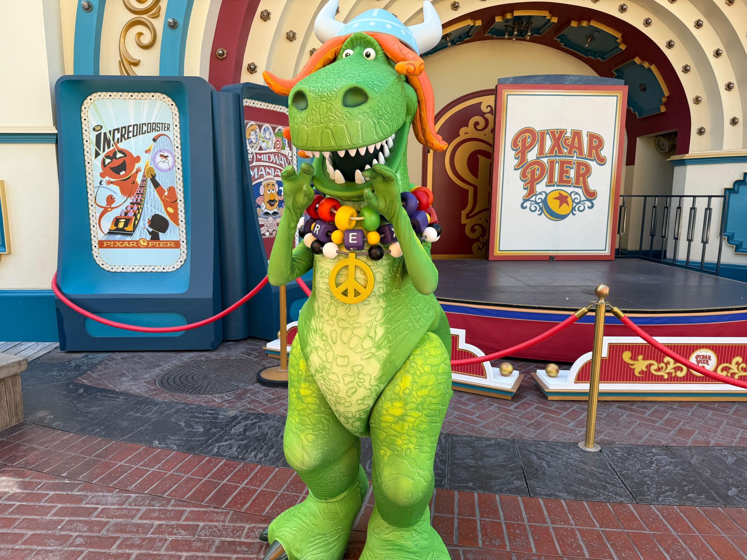 Rex the dinosaur from Toy Story dressed as a hippie, standing in front of a Pixar Pier stage at a theme park.