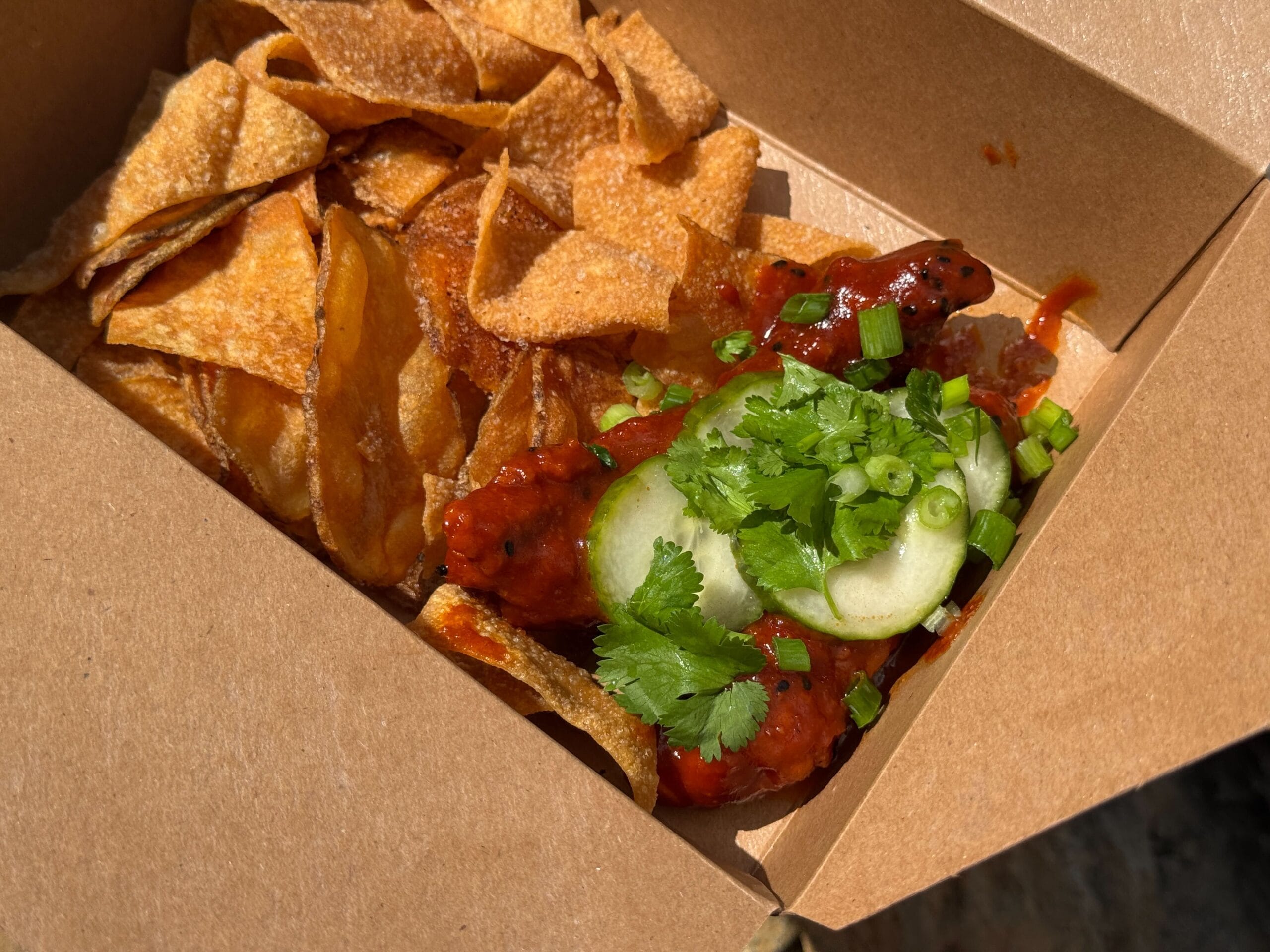 A takeout box at a Disney park filled with crispy chips and gochujang chicken, topped with cucumber, cilantro, and green onions.