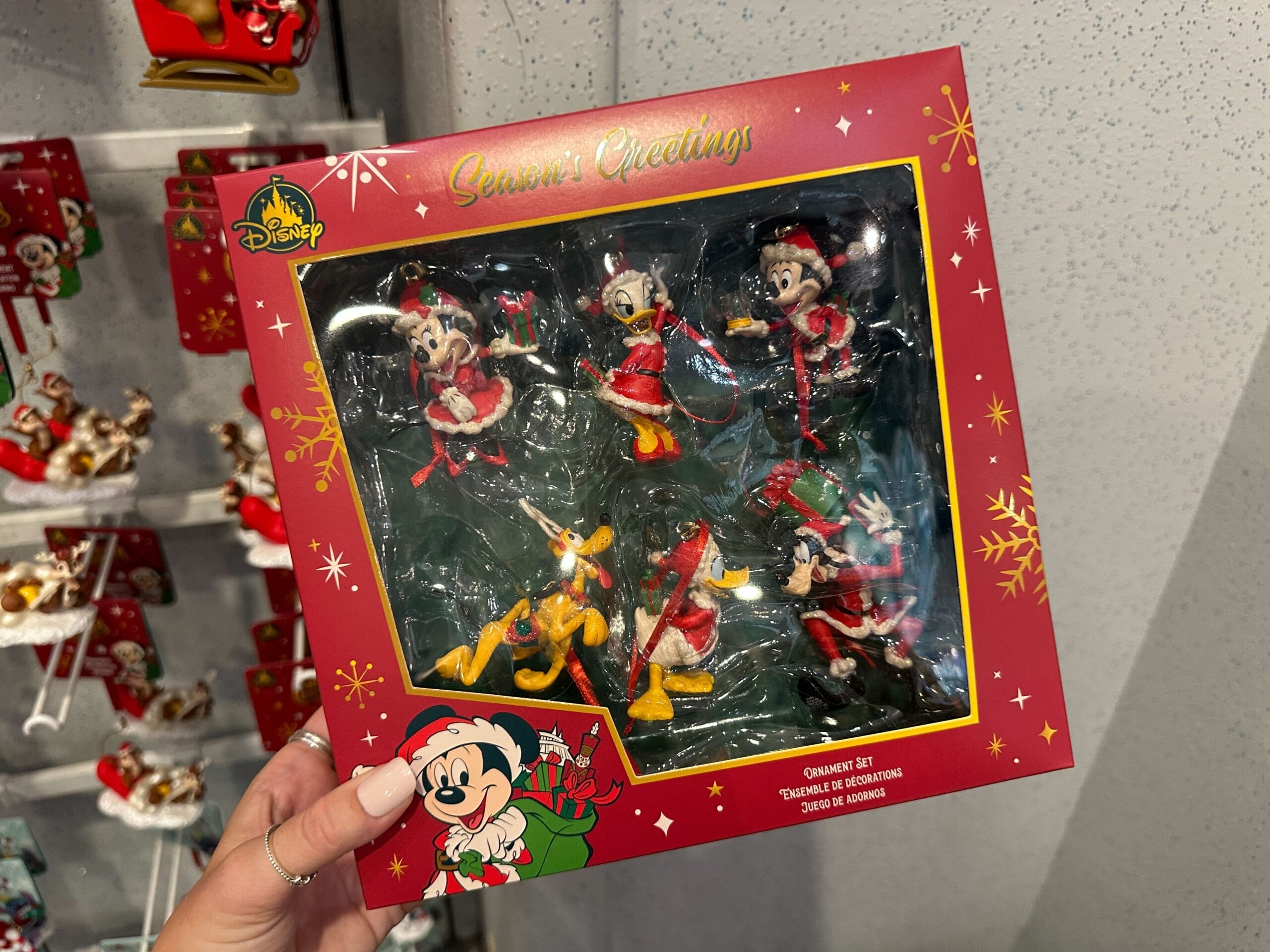 A hand displays Walt Disney World Christmas 2025 ornaments, featuring six characters in holiday attire. No castle is pictured.