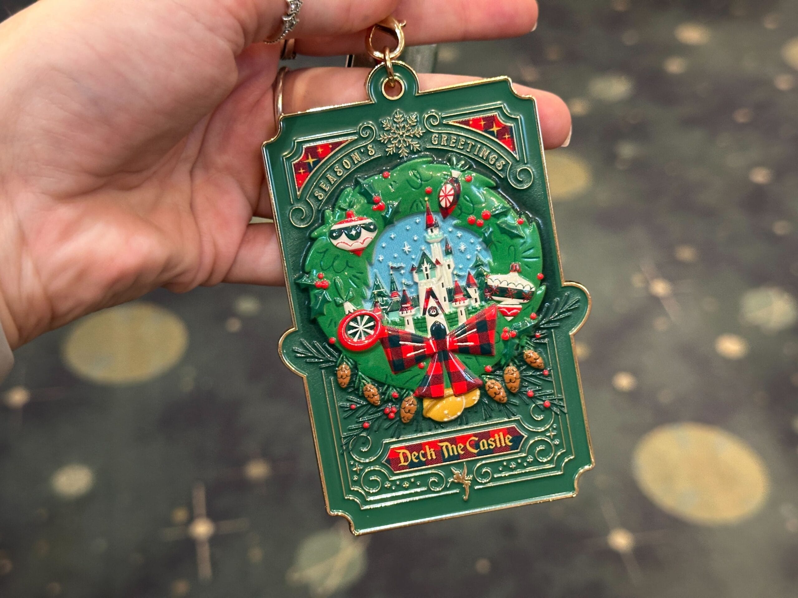 A hand holds a Walt Disney World 2025 "Deck The Castle" ornament featuring wreaths and "Season's Greetings," styled after Cinderella Castle.