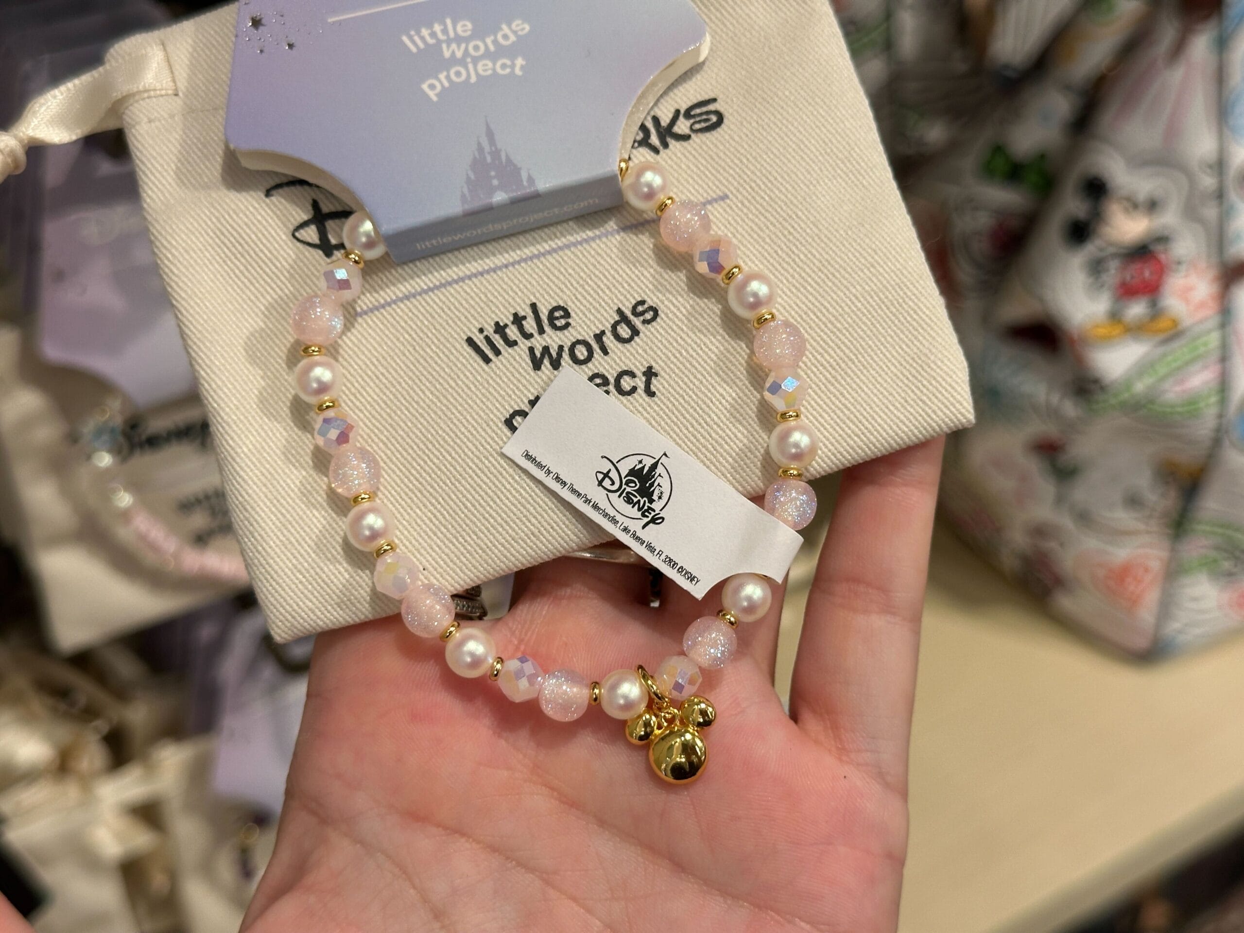 A hand holds a Disney Parks Little Words Project bracelet with iridescent white and pearl beads and a gold Mickey head charm.