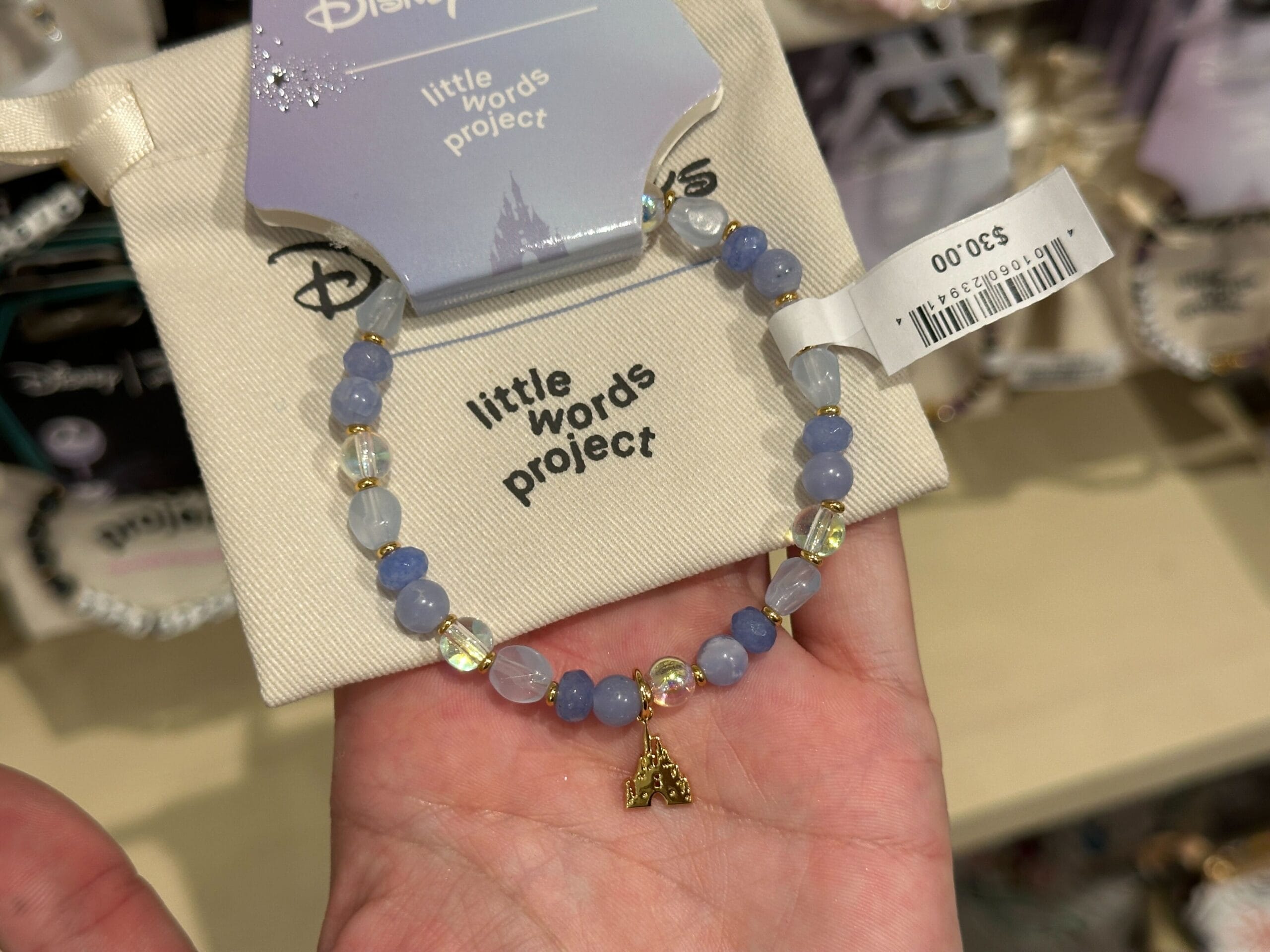 A hand holds a Disney Parks Little Words Project bracelet with iridescent clear and blue beads and a gold Cinderella Castle charm.