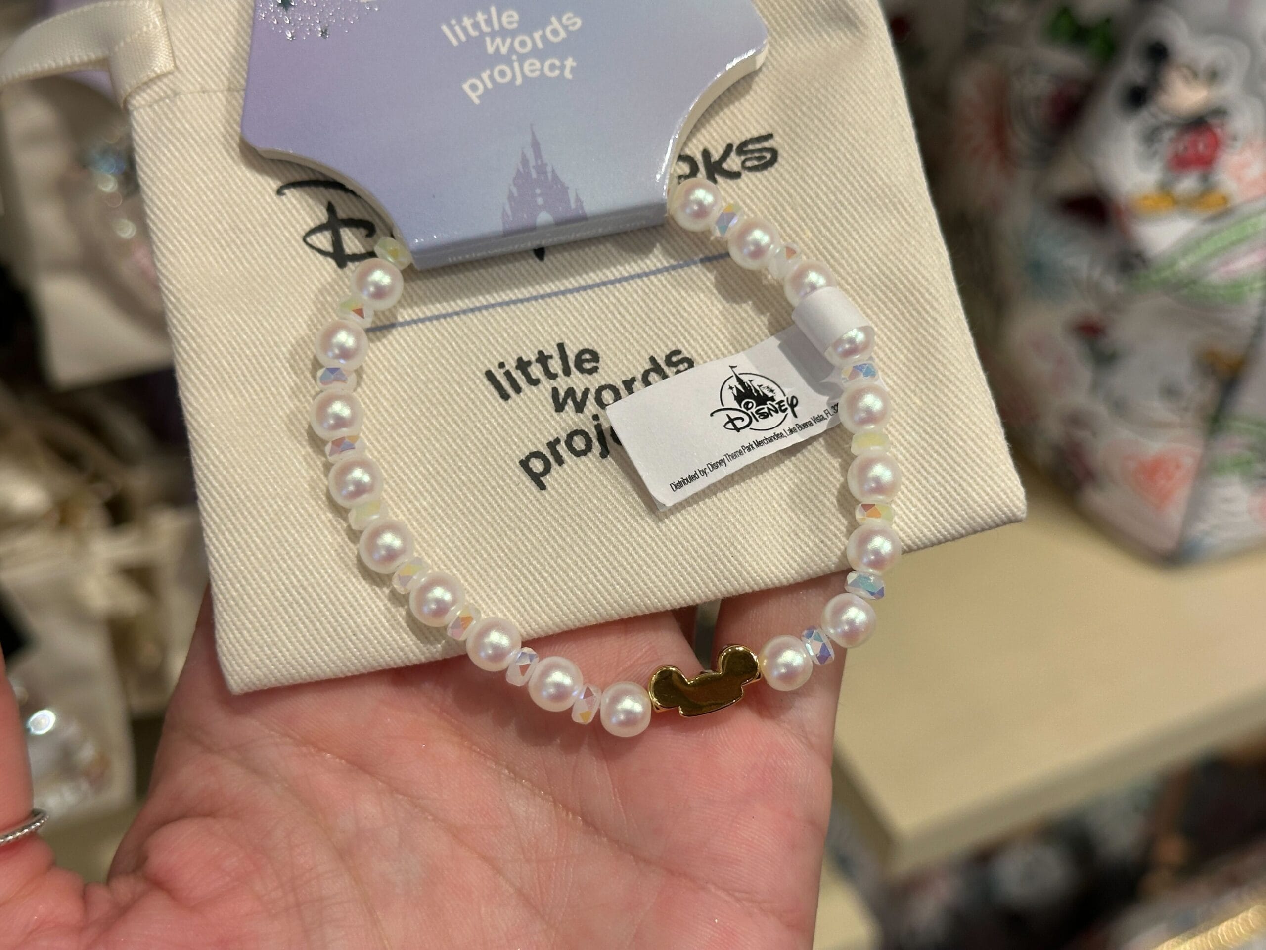 A hand holds a Disney Parks Little Words Project bracelet with iridescent white and pearl beads and a gold Mickey ear hat charm.