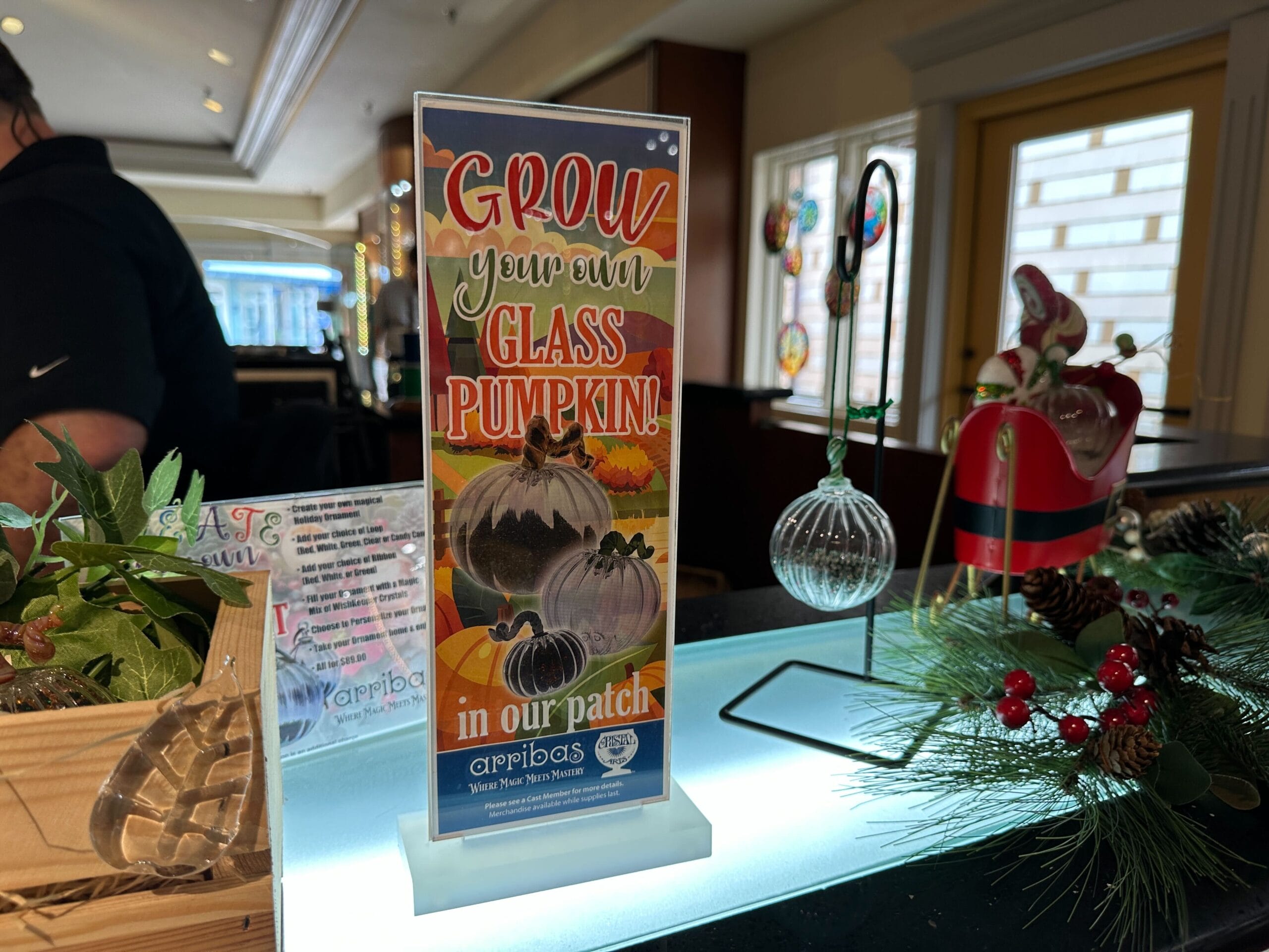 A festive sign at a decorated counter says "Grow your own glass pumpkin! in our patch" on the counter at Crystal Arts in Walt Disney World.
