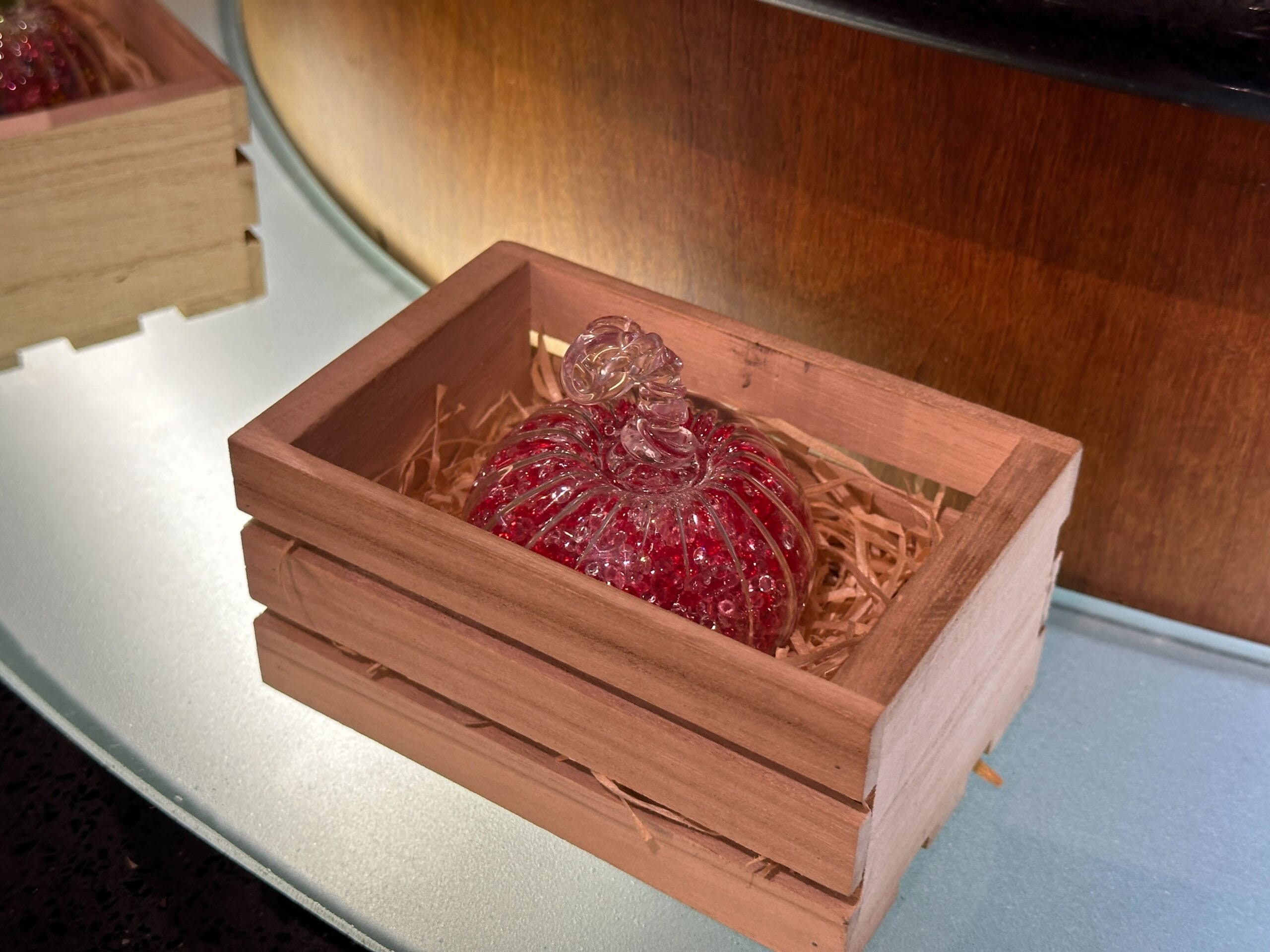 A glass pumpkin with red details rests in straw inside a small wooden crate, resembling festive fall decor at Disney parks.