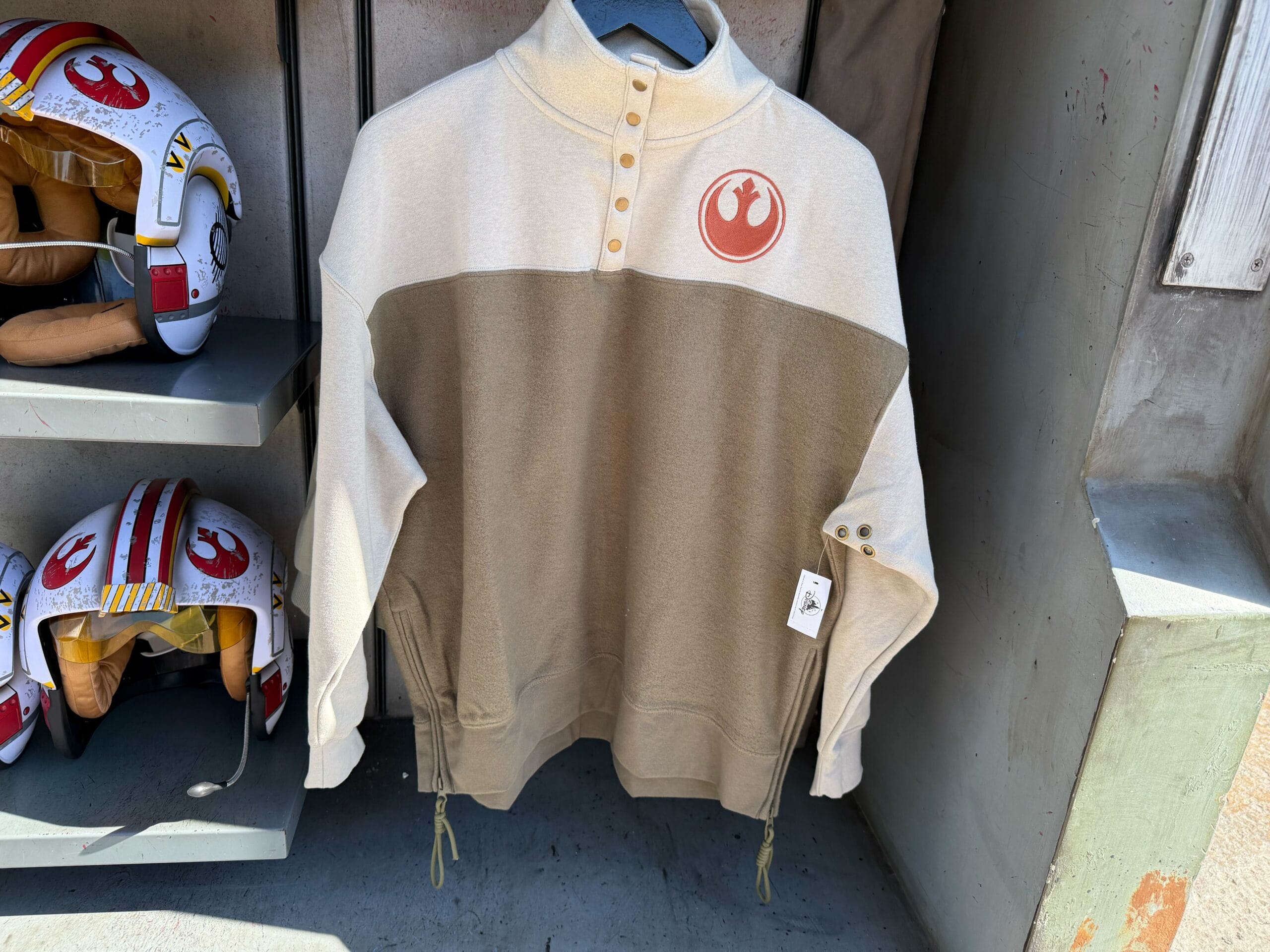 A beige and olive Star Wars merch pullover with a red Rebel Alliance logo, displayed by two pilot helmets.