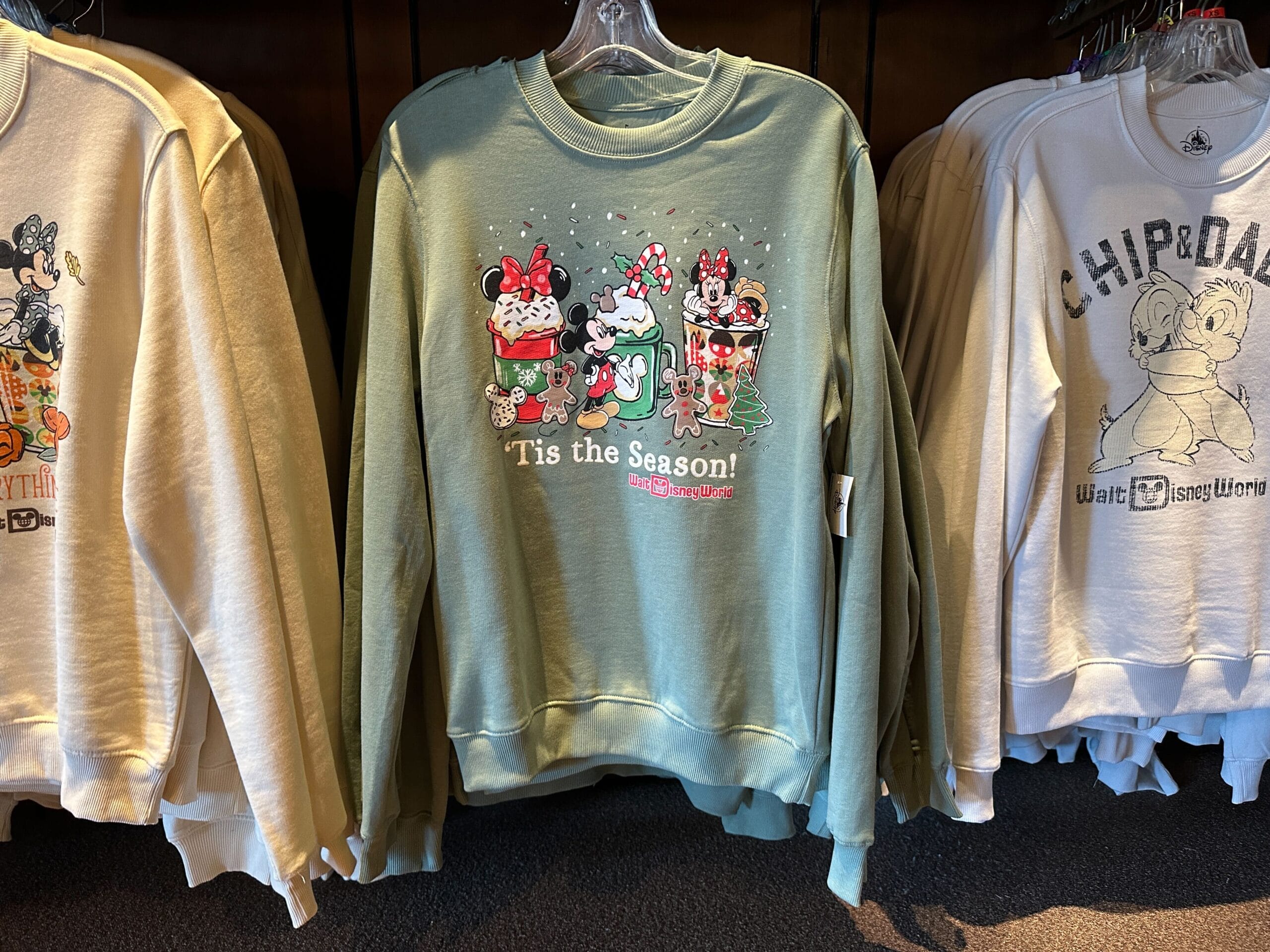Festive green sweatshirt with Walt Disney World characters and “‘Tis the Season!” on a merch rack, evoking park holiday spirit.