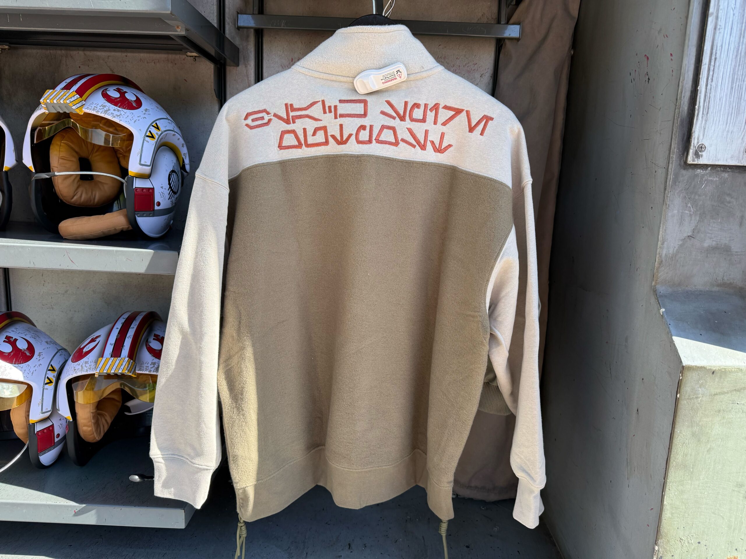 A beige and tan jacket with red Star Wars Aurebesh text, seen at Galaxy's Edge near helmet display shelves.