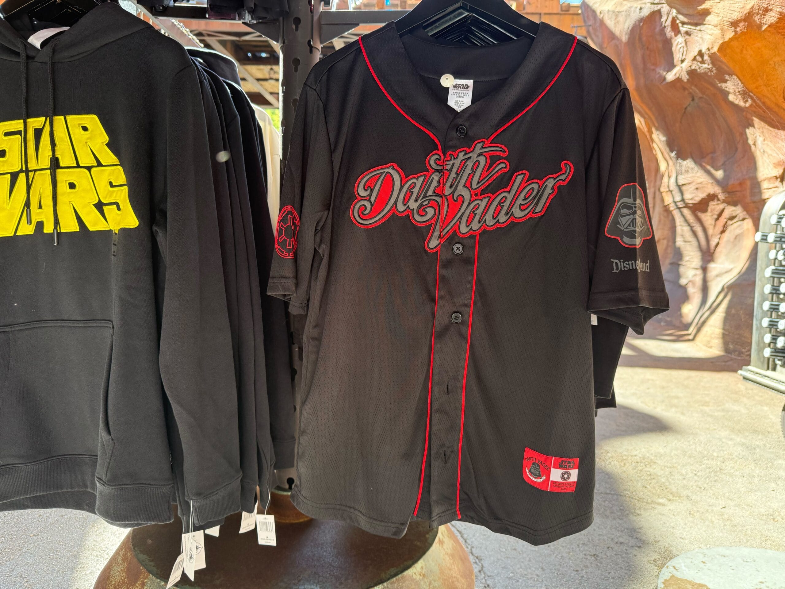Black baseball jersey with red trim and "Darth Vader" hangs by a black Star Wars hoodie—perfect Star Wars merch for fans.