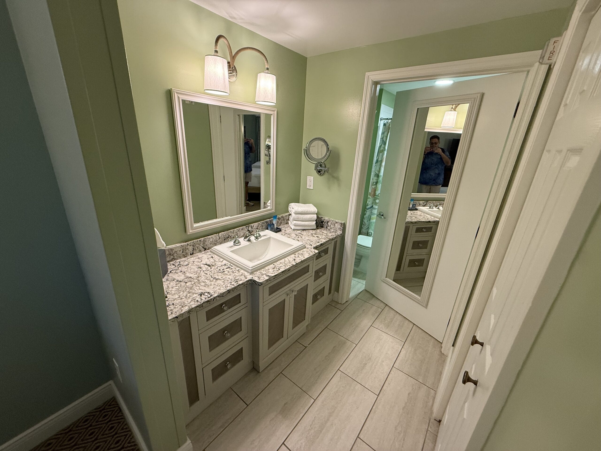 Bathroom at Disney's Vero Beach Resort features granite countertop, sink, mirrors, green walls; person visible in mirror.