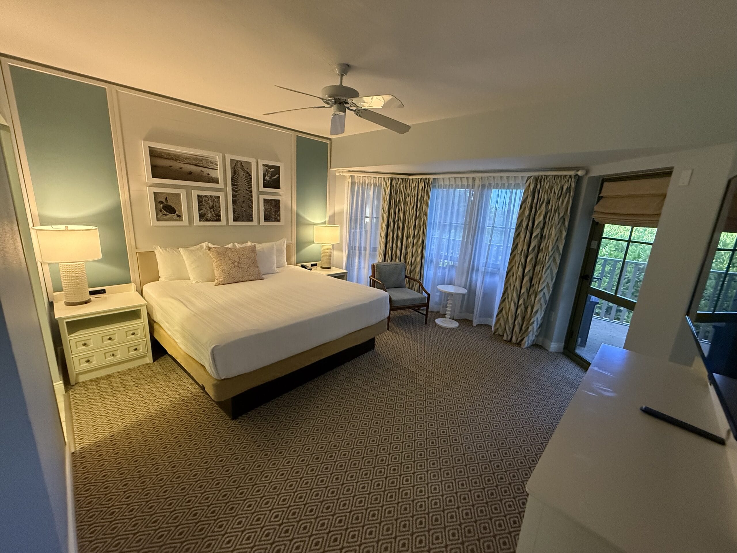 A cozy, Disney-inspired cottage hotel room with a king bed, two chairs by bright windows, and warm, fairytale-like lighting.