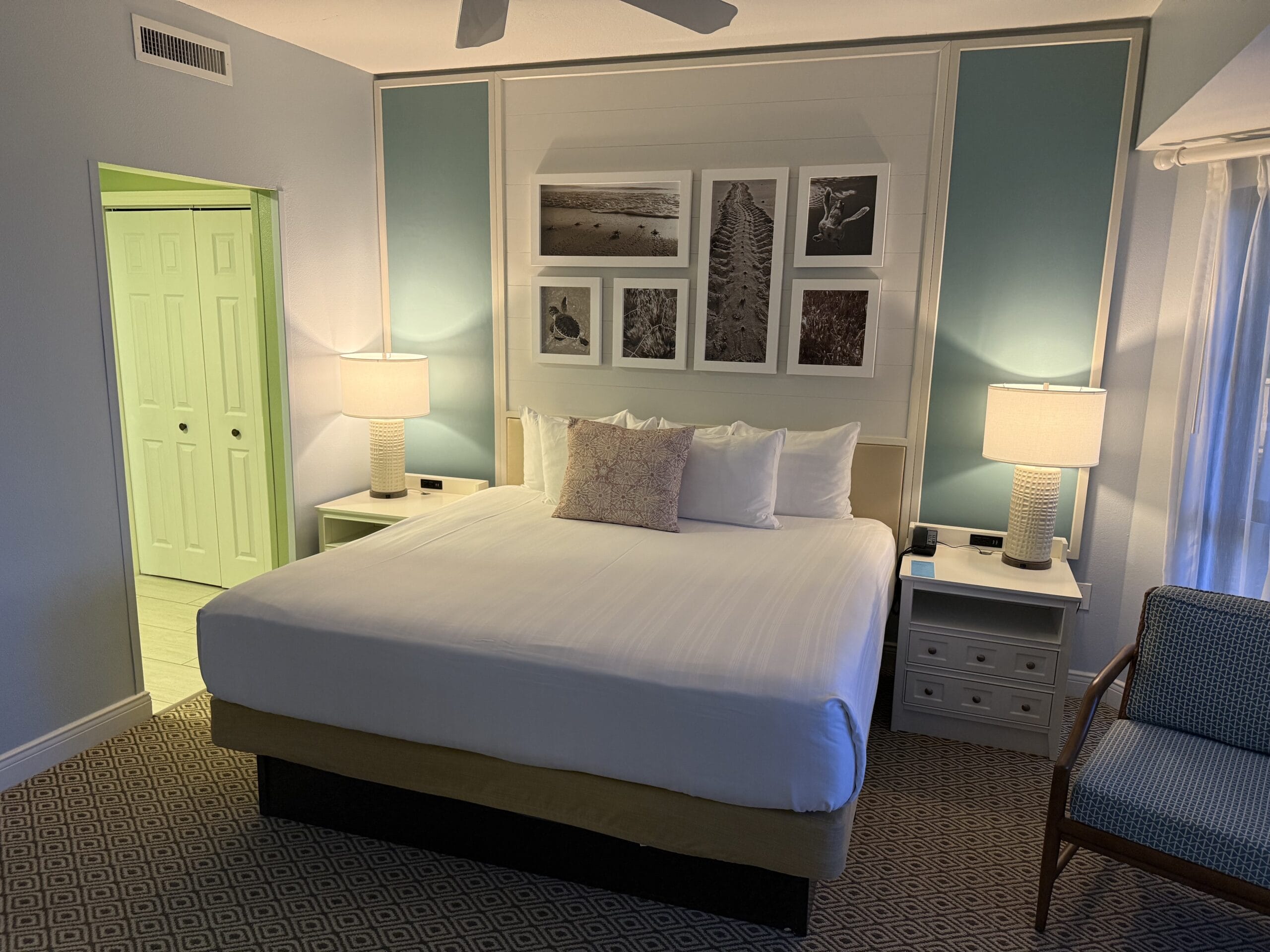 A serene Disney's Vero Beach Resort room with king bed, two lamps, Disney artwork, and a cozy chair near a sunlit window.