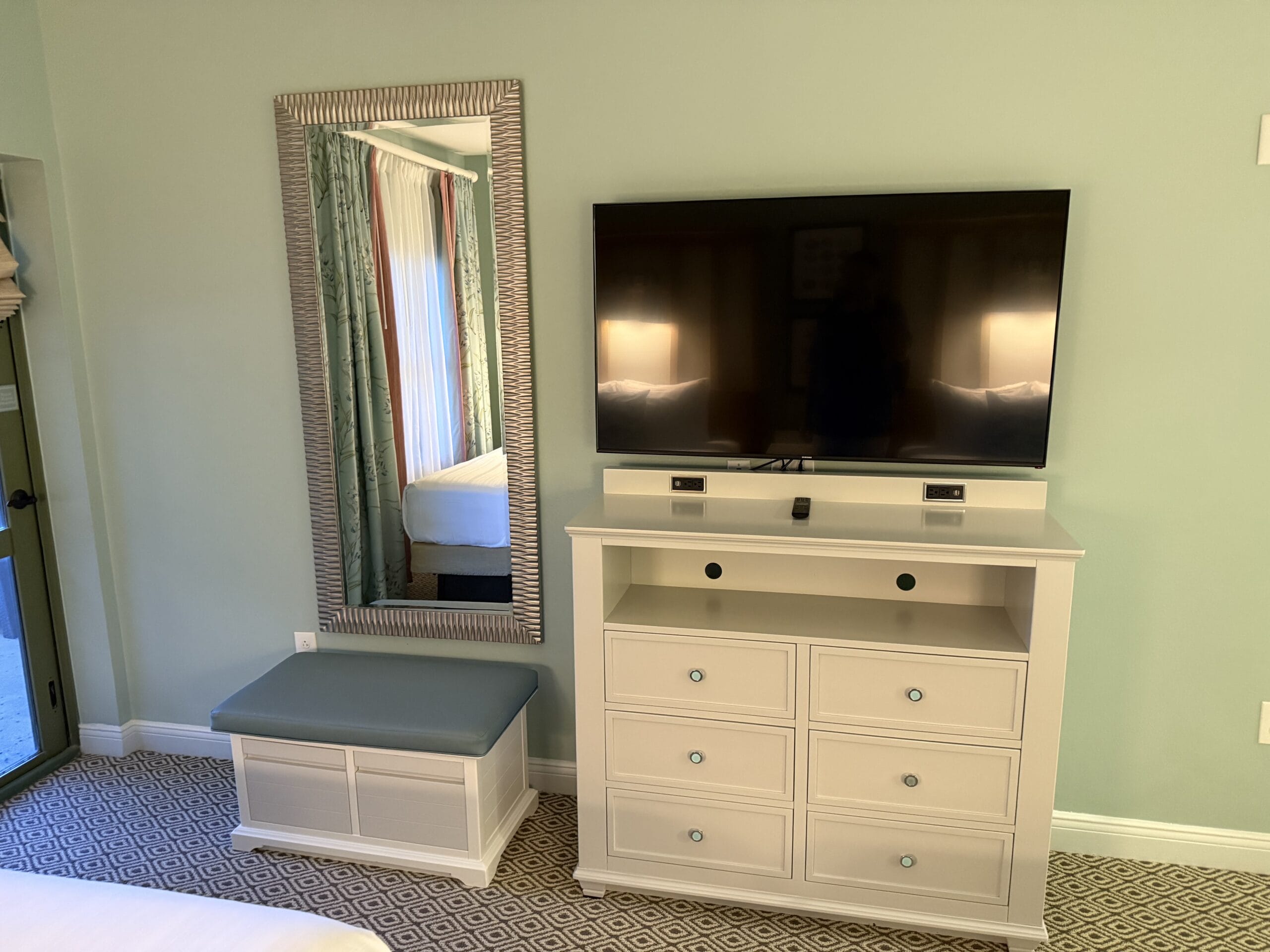 A flat-screen TV on a white dresser next to a bench and tall mirror in a cozy cottage at Disney’s Vero Beach Resort, not a castle room.