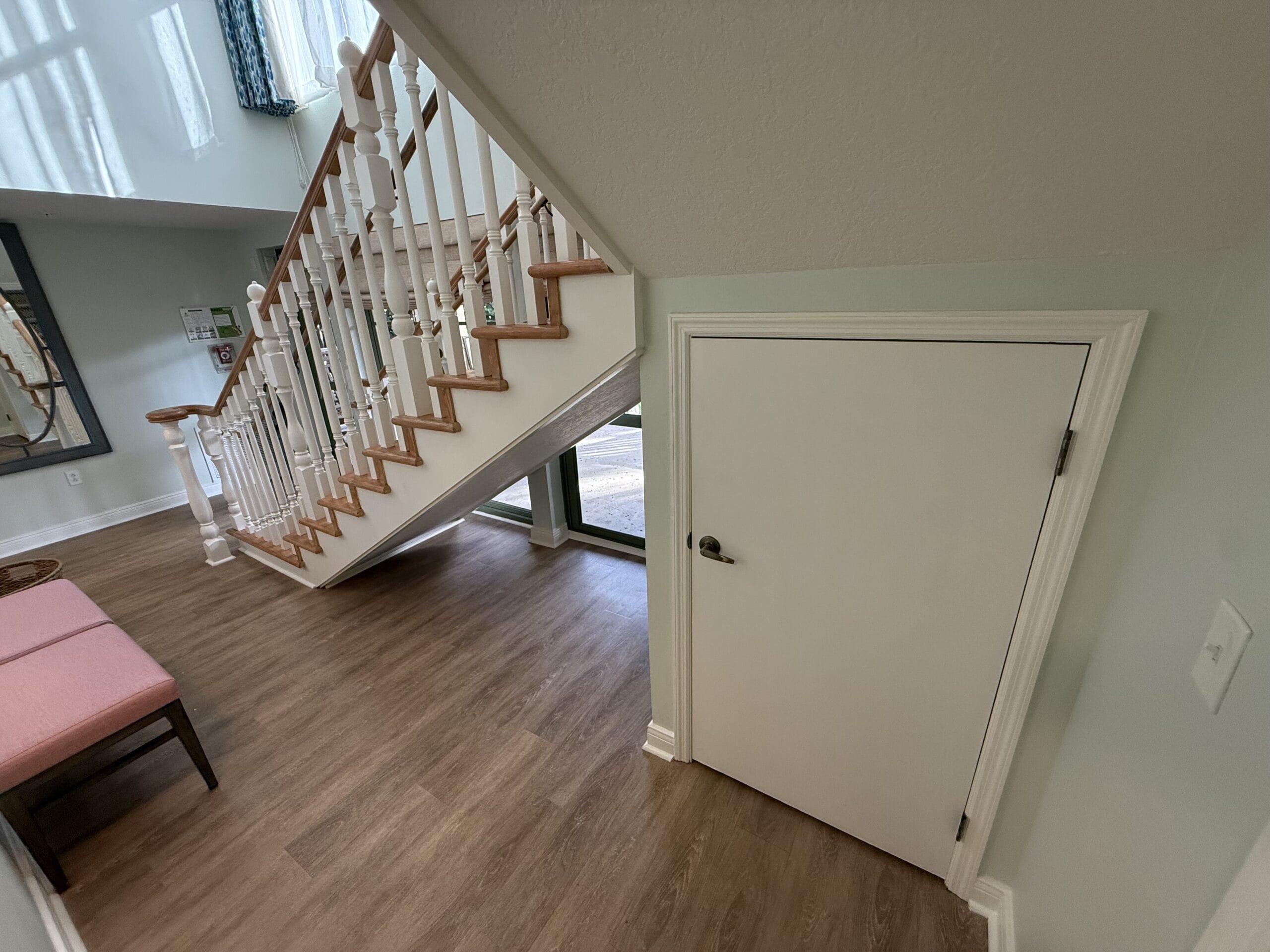 A charming white door beneath stairs in a Disney Resort-style home, with wood floors and a pink bench evoking storybook magic.