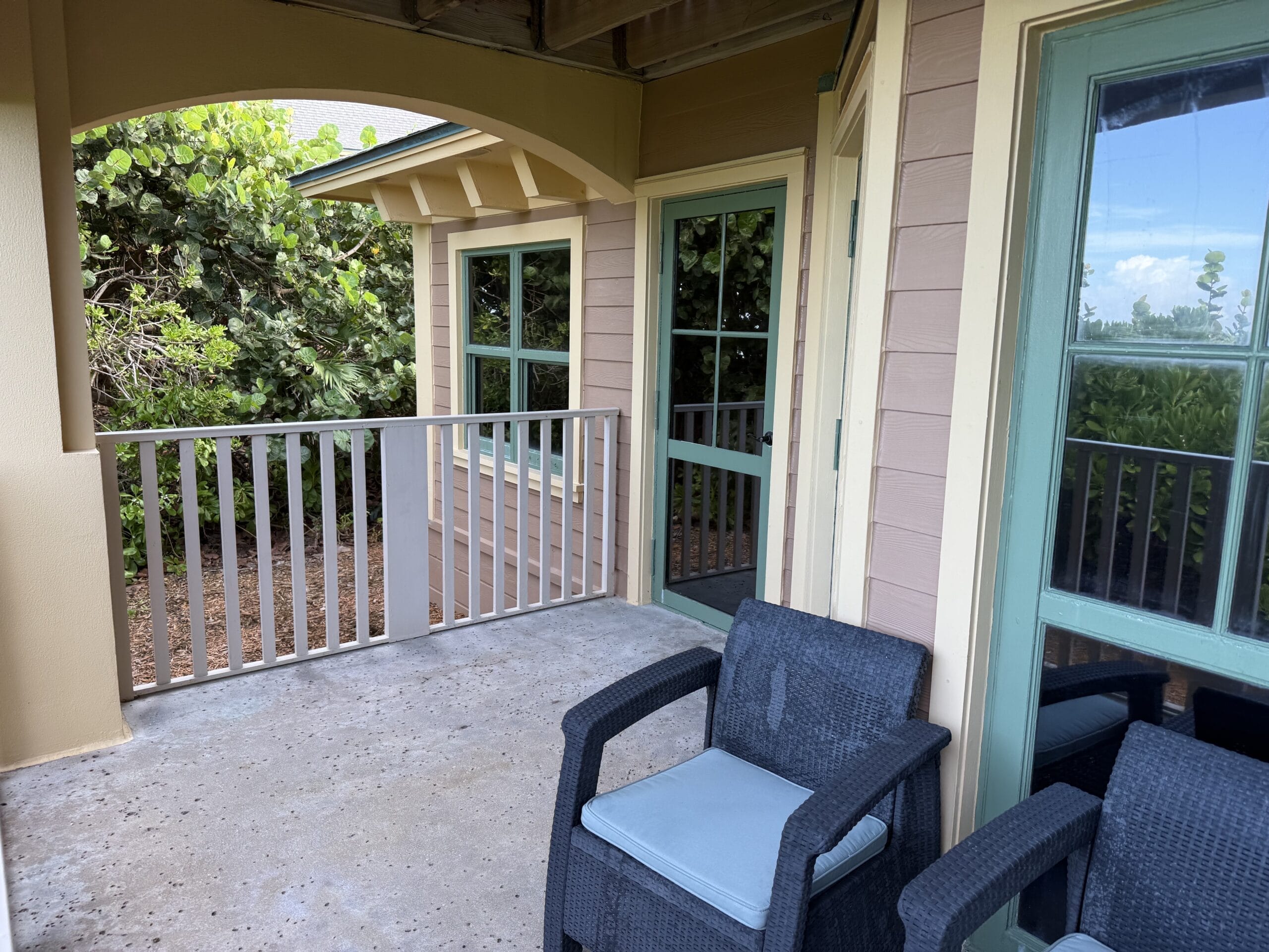 A cozy porch at Disney’s Vero Beach Resort features black chairs, blue cushions, green-trimmed doors, and lush garden views.