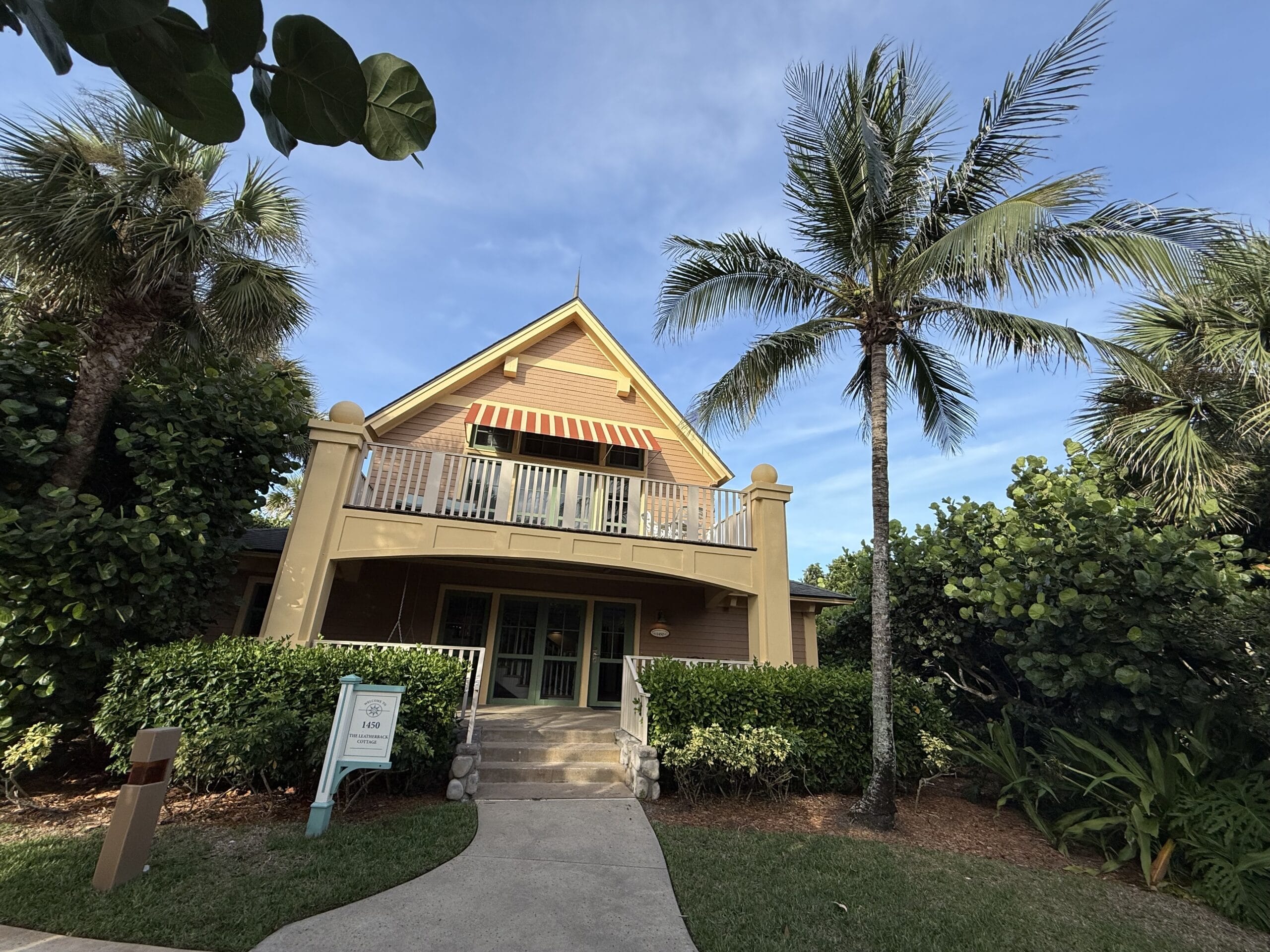 A yellow cottage with a balcony, palm trees, and lush greenery, evoking Disney's Vero Beach Resort on a clear blue sky day.