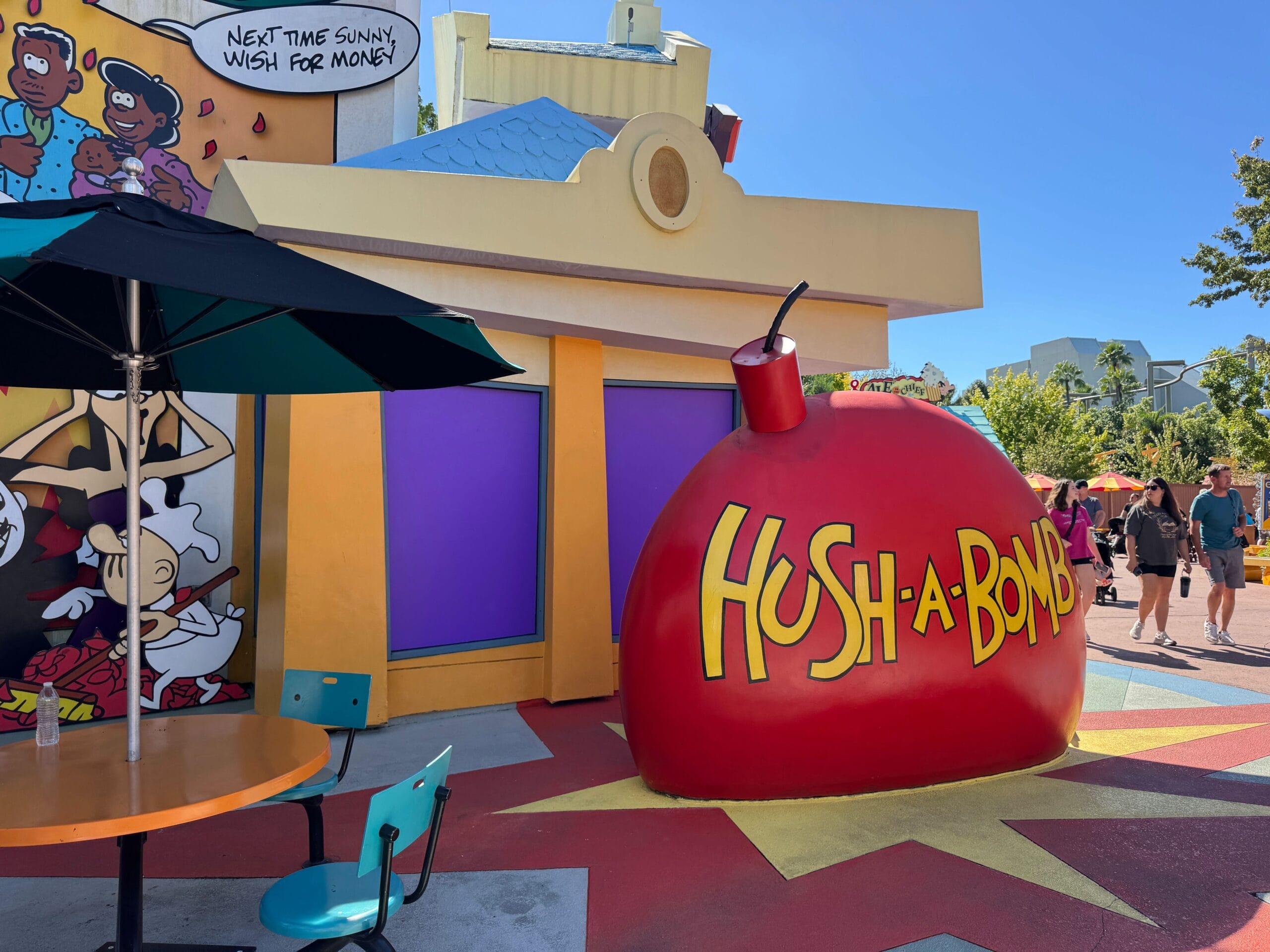 Large red cartoony "HUSH-A-BOMB" prop sits outside the Wossamotta-U gift shop in Universal Islands of Adventure. Solid purple panels cover the windows.