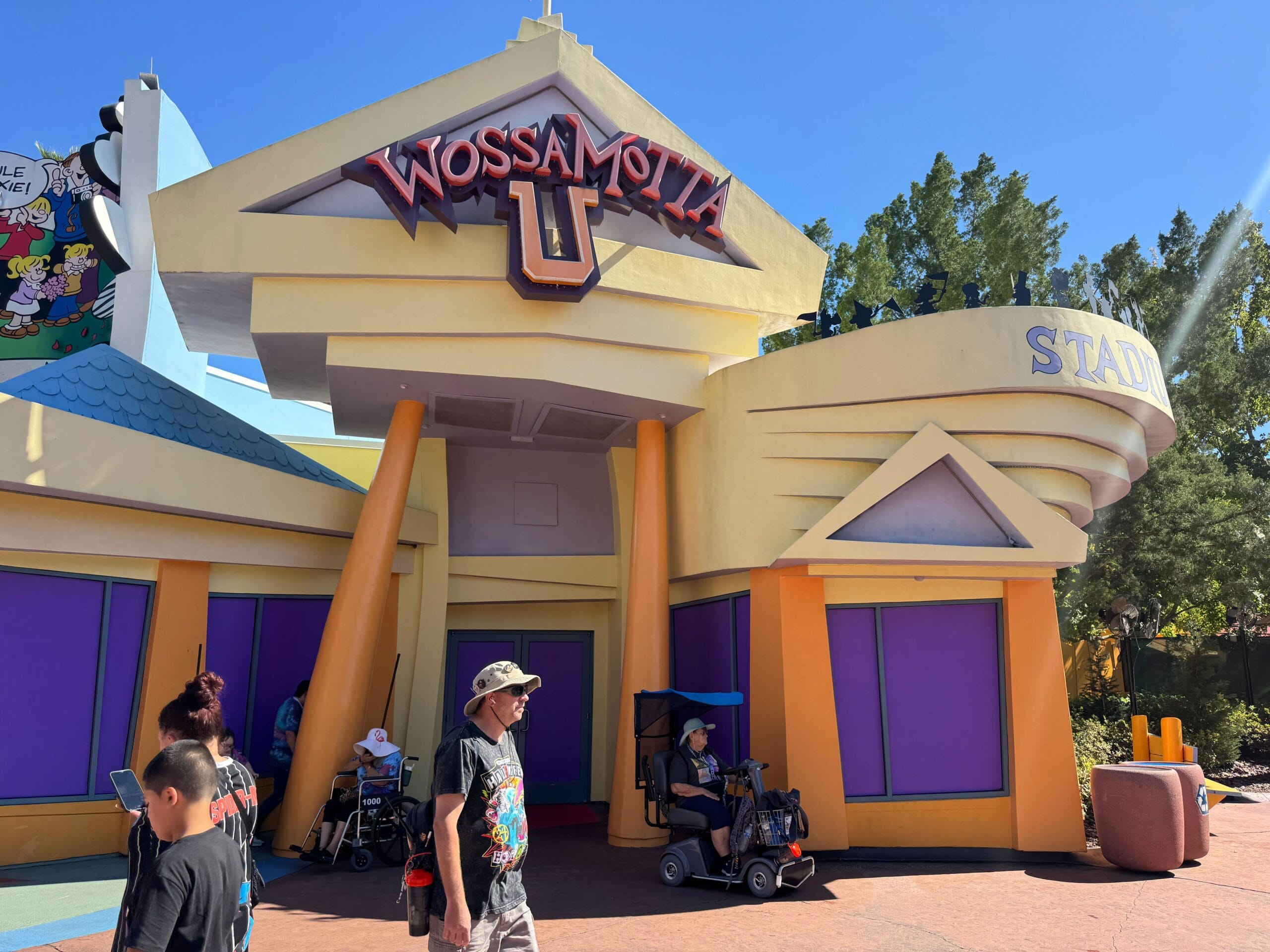 People relax in the shade outside the vibrant, cartoon-inspired exterior of the Wossamotta-U gift shop in Universal Islands of Adventure. Solid purple panels cover the windows and doors.