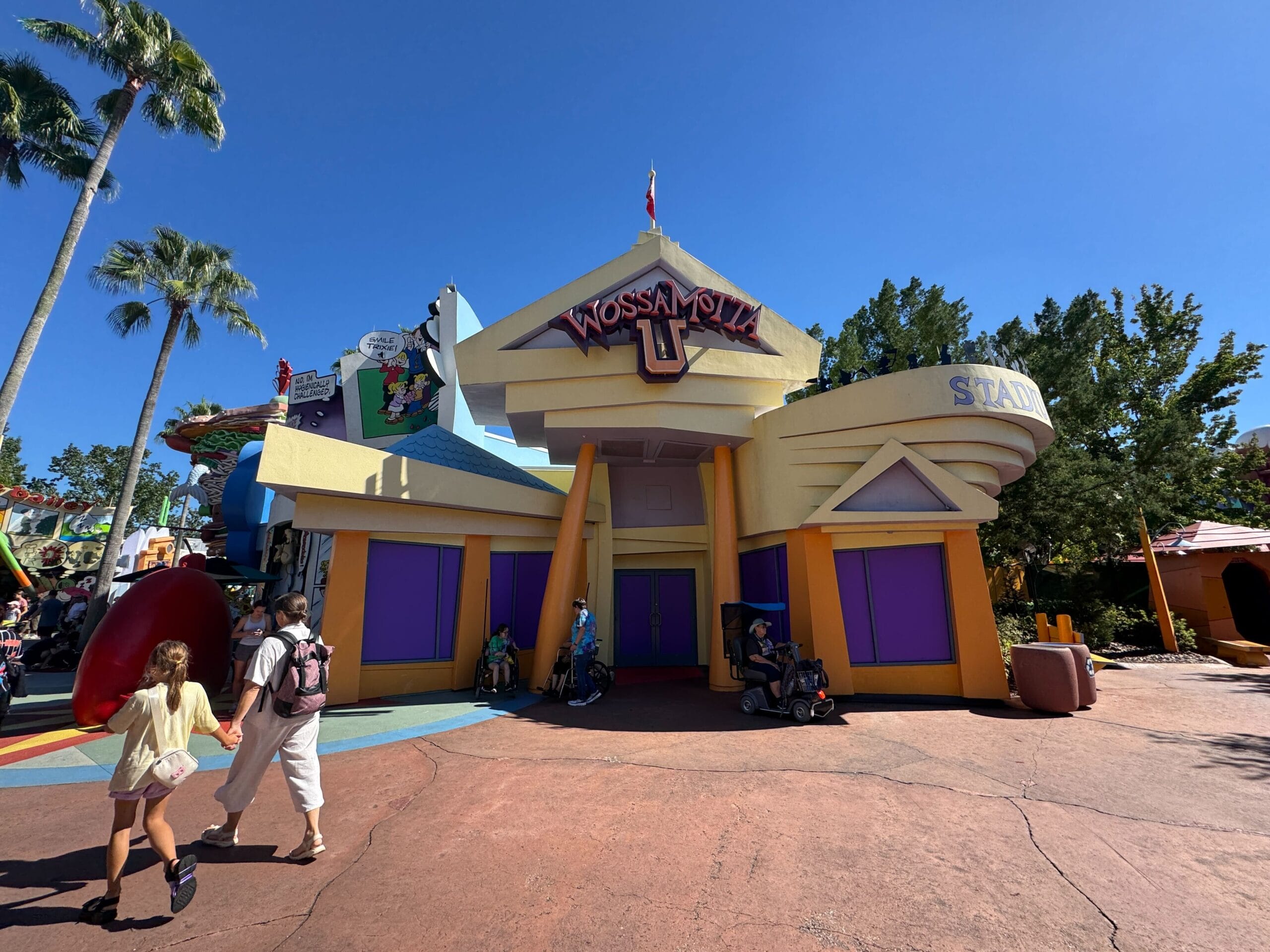 People relax in the shade outside the vibrant, cartoon-inspired exterior of the Wossamotta-U gift shop in Universal Islands of Adventure. Solid purple panels cover the windows and doors.