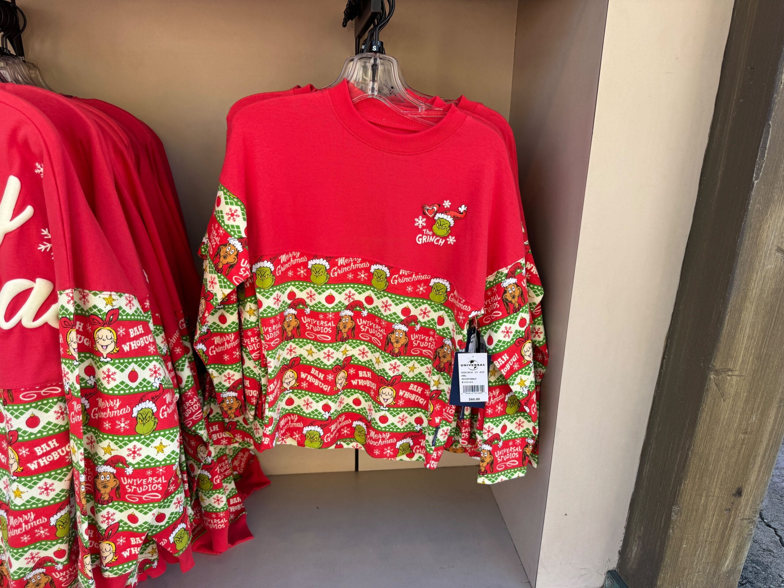 Red and green Grinchmas sweater with "The Grinch" and festive holiday patterns, displayed on a rack at Universal Orlando, not Disney.