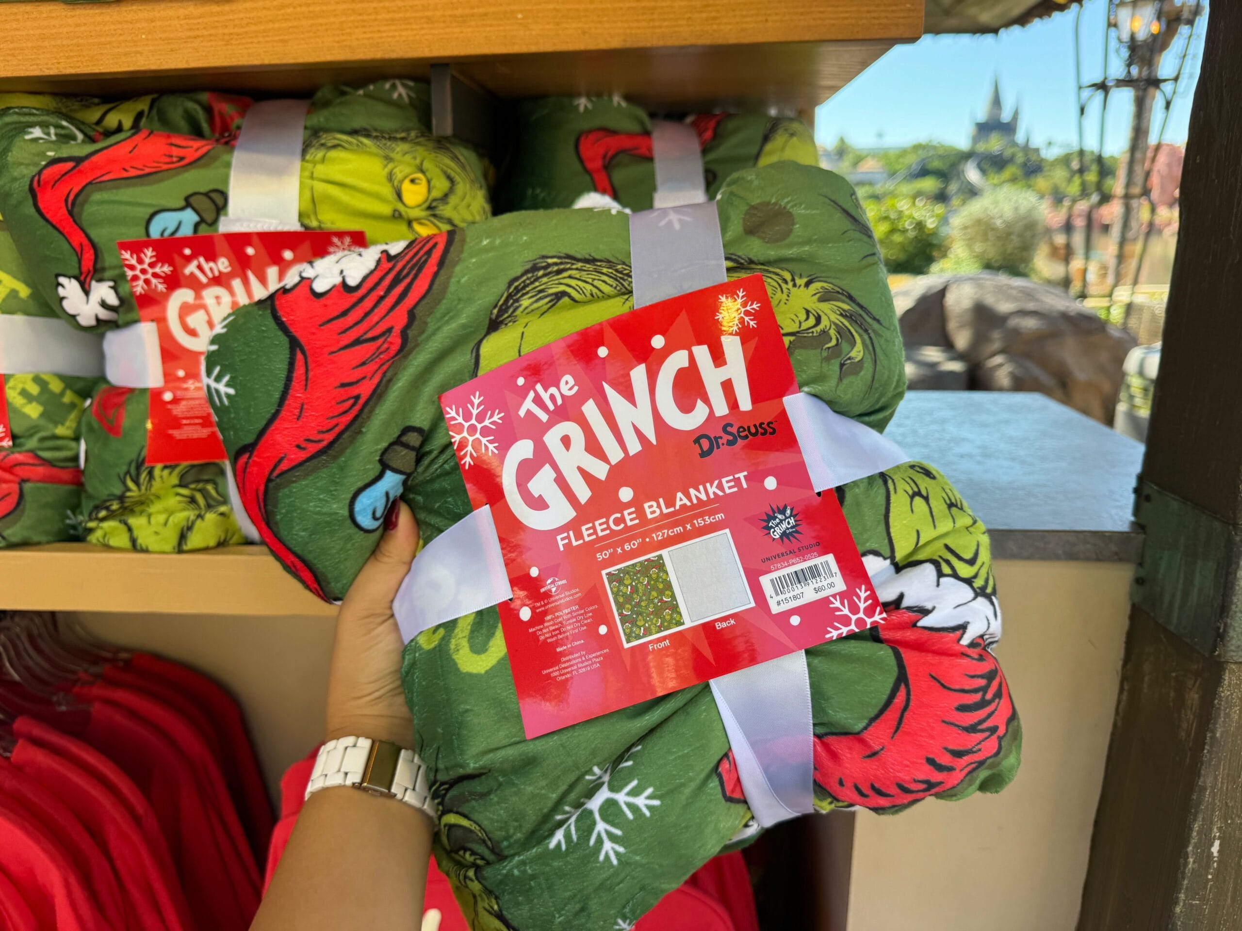 Hand holding Grinchmas fleece blanket with red label, displayed at Universal Orlando—no Disney castle like those in Disneyland parks.