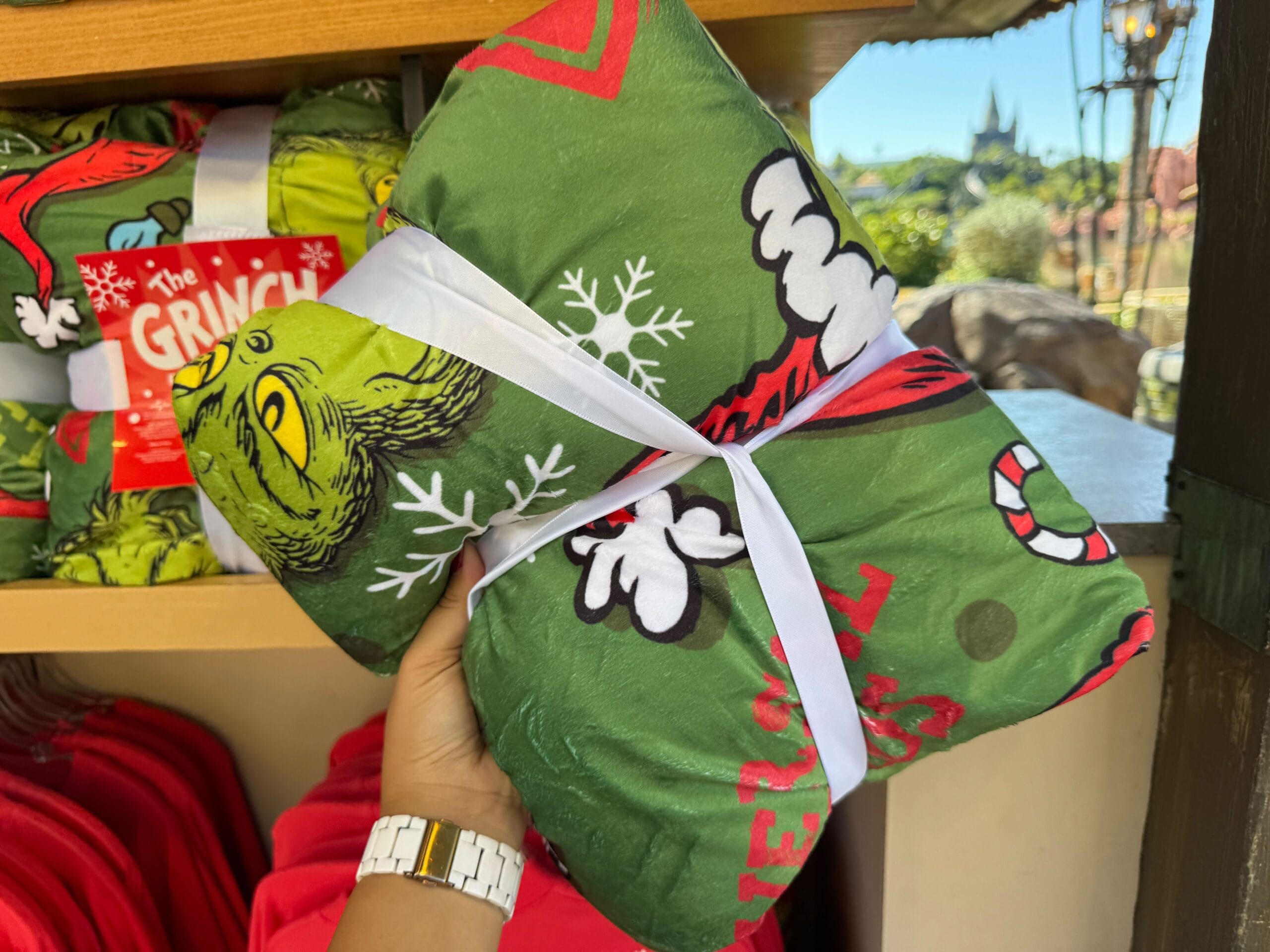 A hand holds Grinchmas merch—a green Grinch-themed blanket tied with a white ribbon—at Universal Orlando, not a Disney park.