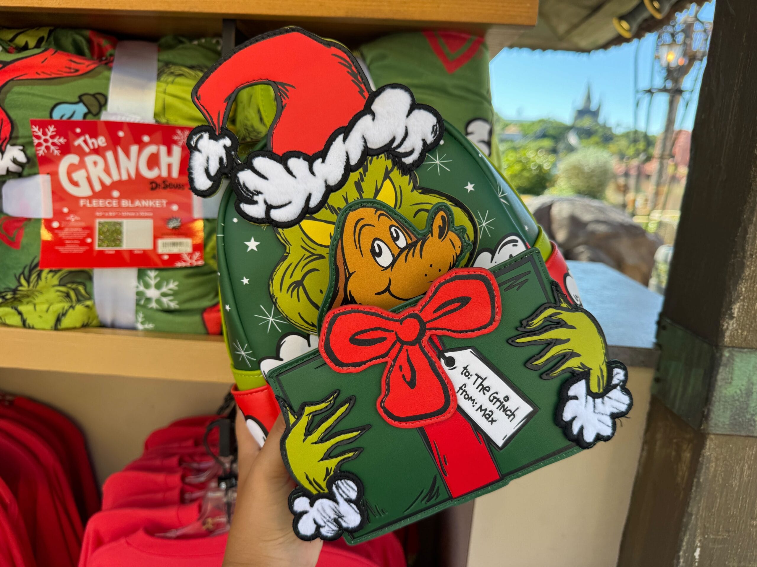 A hand holds Grinchmas merch: a Grinch holiday backpack with a Santa hat and tag, but no Disney castle like at Magic Kingdom.