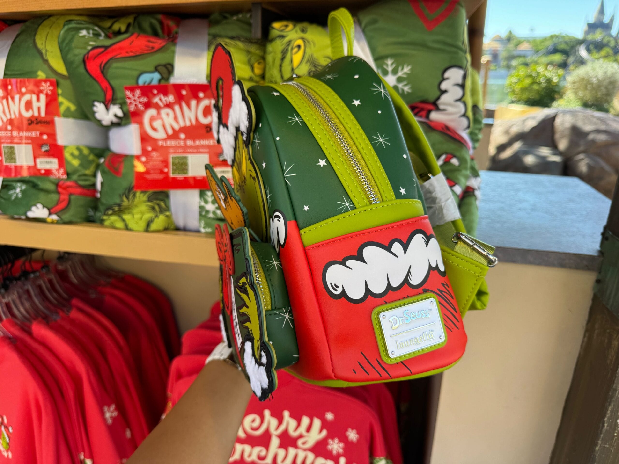 A hand holds a small Grinch-themed backpack in front of Grinchmas merchandise at Universal Orlando, not Disney Parks like Cinderella Castle.