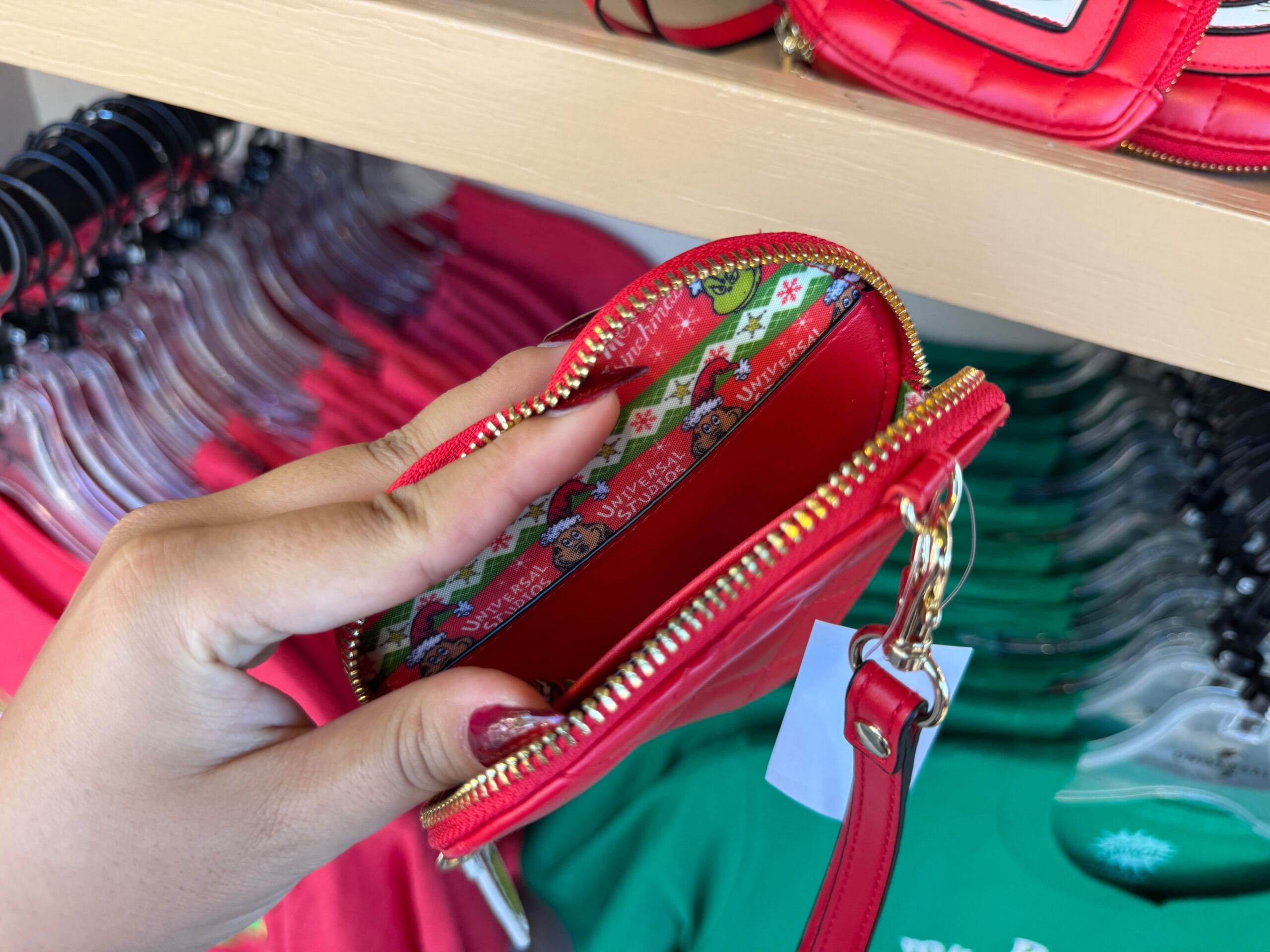 Hand holding a small red zippered pouch with cartoon dog lining, surrounded by Grinchmas merch and colorful shirts—not at Disney Castle.