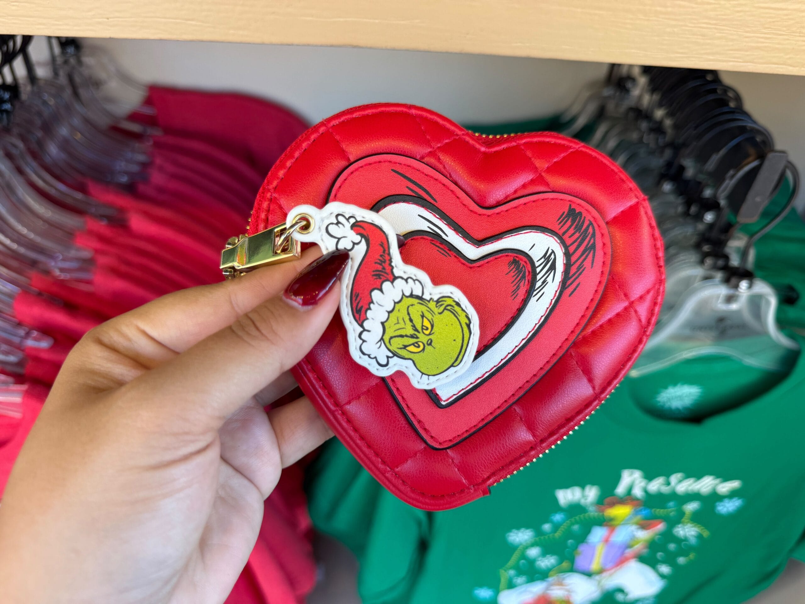 A hand holds a red heart-shaped coin purse with a Grinch tag; festive merch hangs behind, but no Disney castle in this display.
