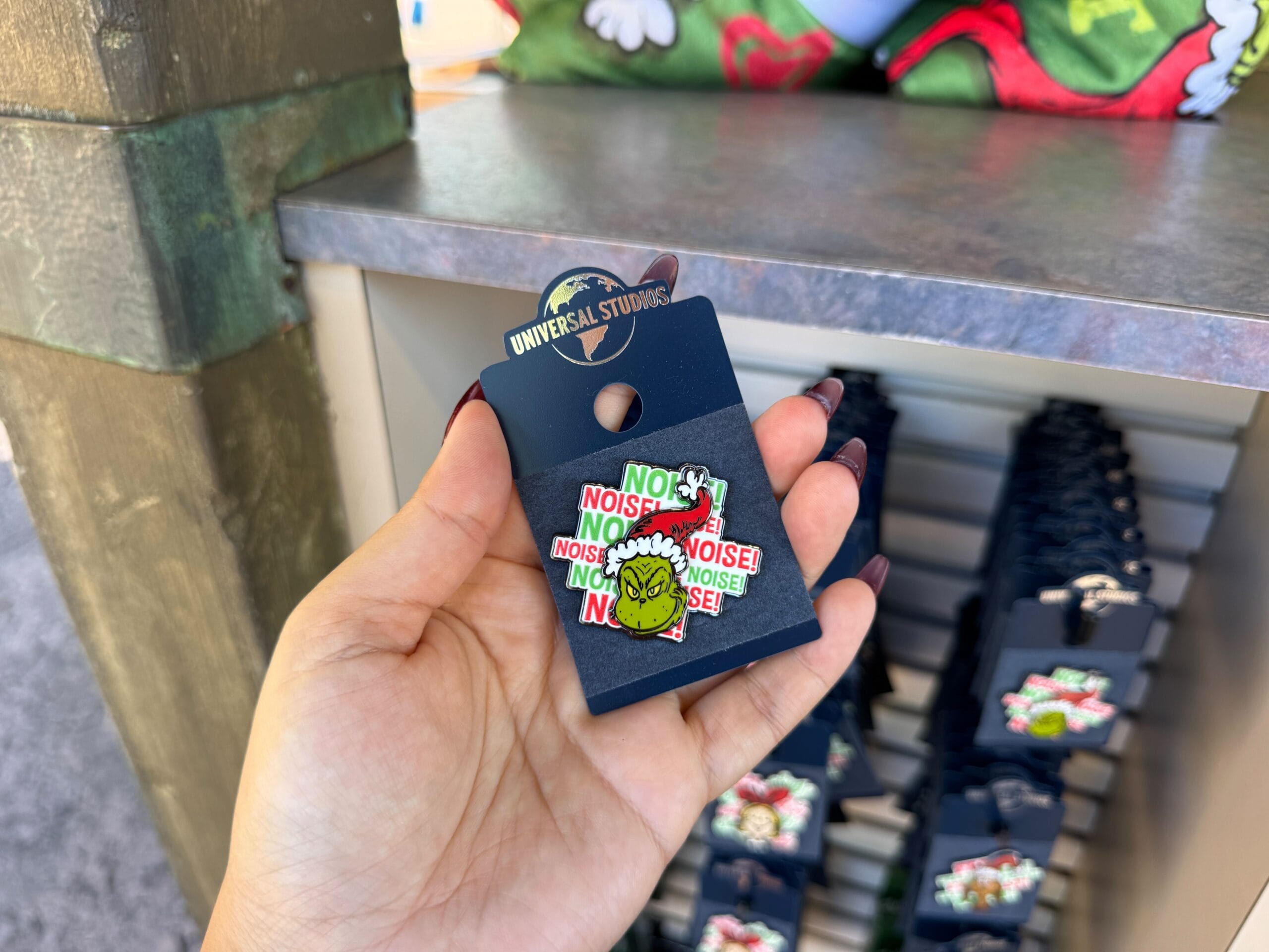 A hand displays a Grinchmas "NOISE! NOISE! NOISE!" pin at Universal Orlando—no Cinderella or Sleeping Beauty Castle in sight here.
