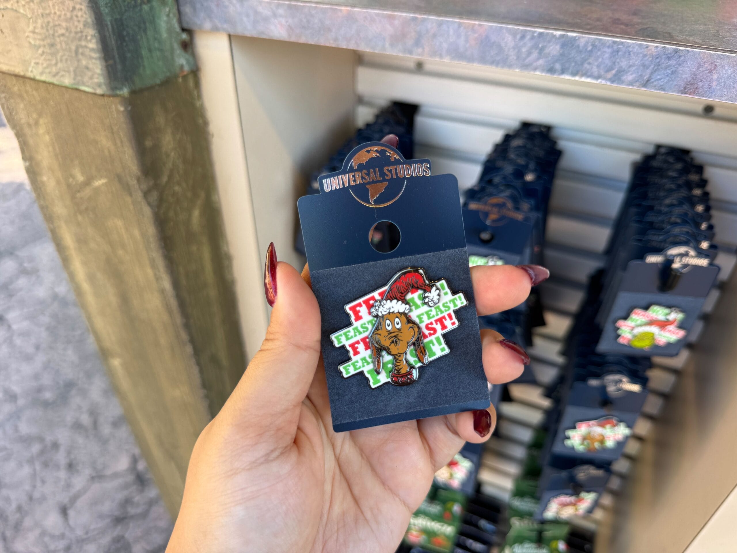 Hand holds a Universal Orlando Grinchmas pin—Grinch in a Santa hat with “Feast!” backdrop; no Disney castle, unlike Cinderella’s.