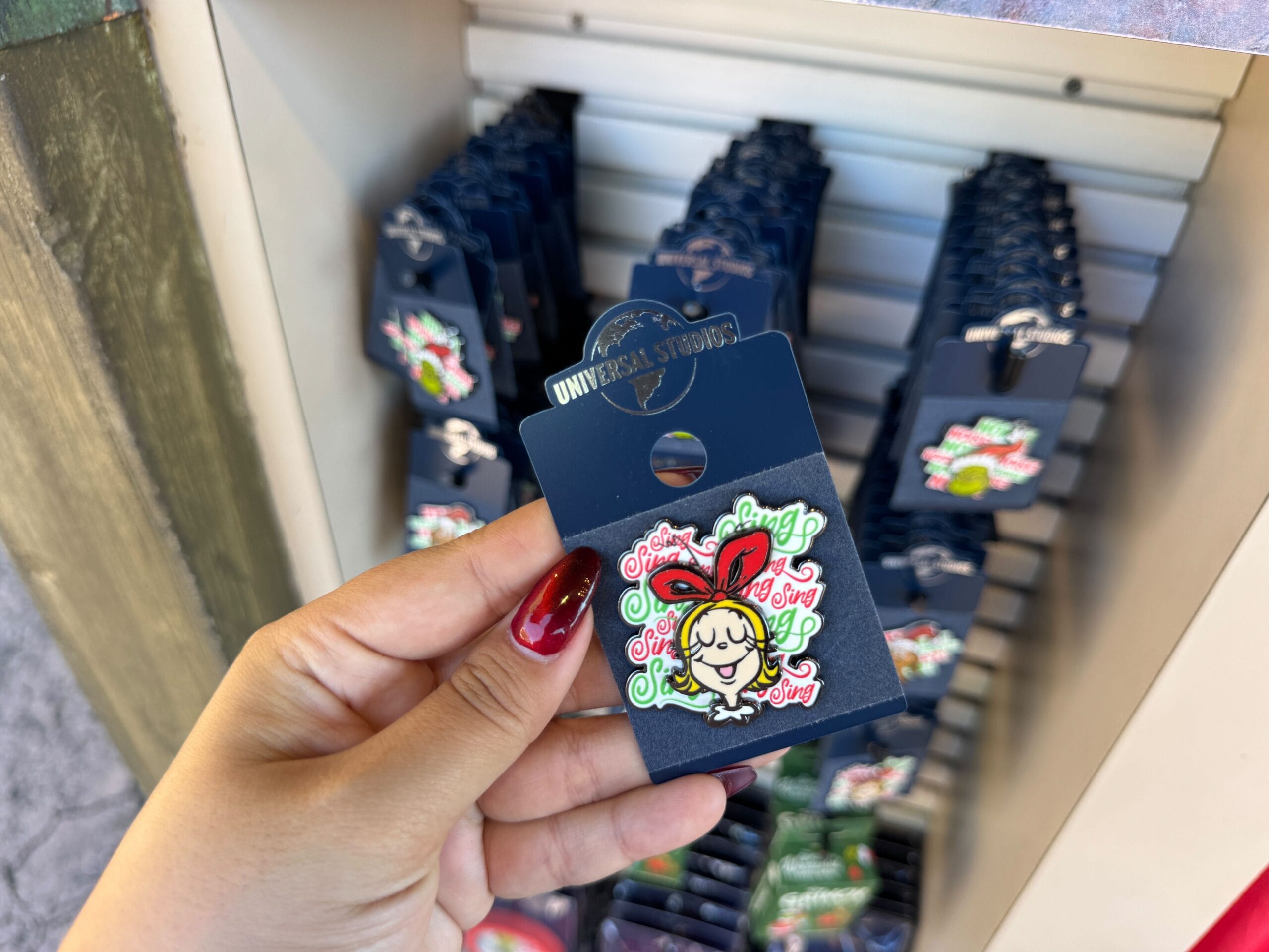 A hand holds a Universal Orlando pin of a cartoon girl in a red bow, similar in style to Disney pins, with more pins behind.