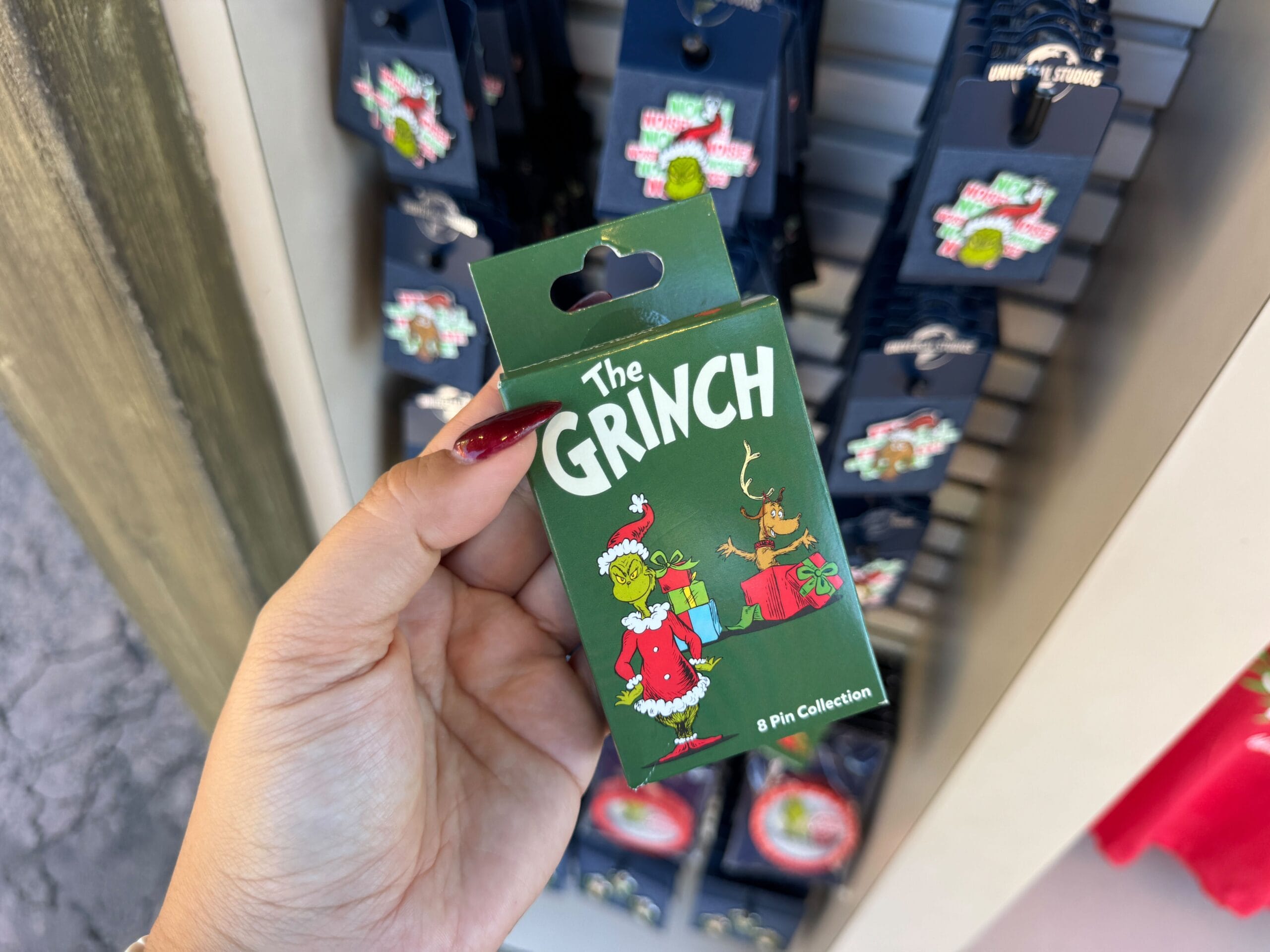 Hand holding a Grinchmas pin collection in front of a Universal Orlando Grinch display, not a Disney castle like Cinderella’s or Sleeping Beauty’s.