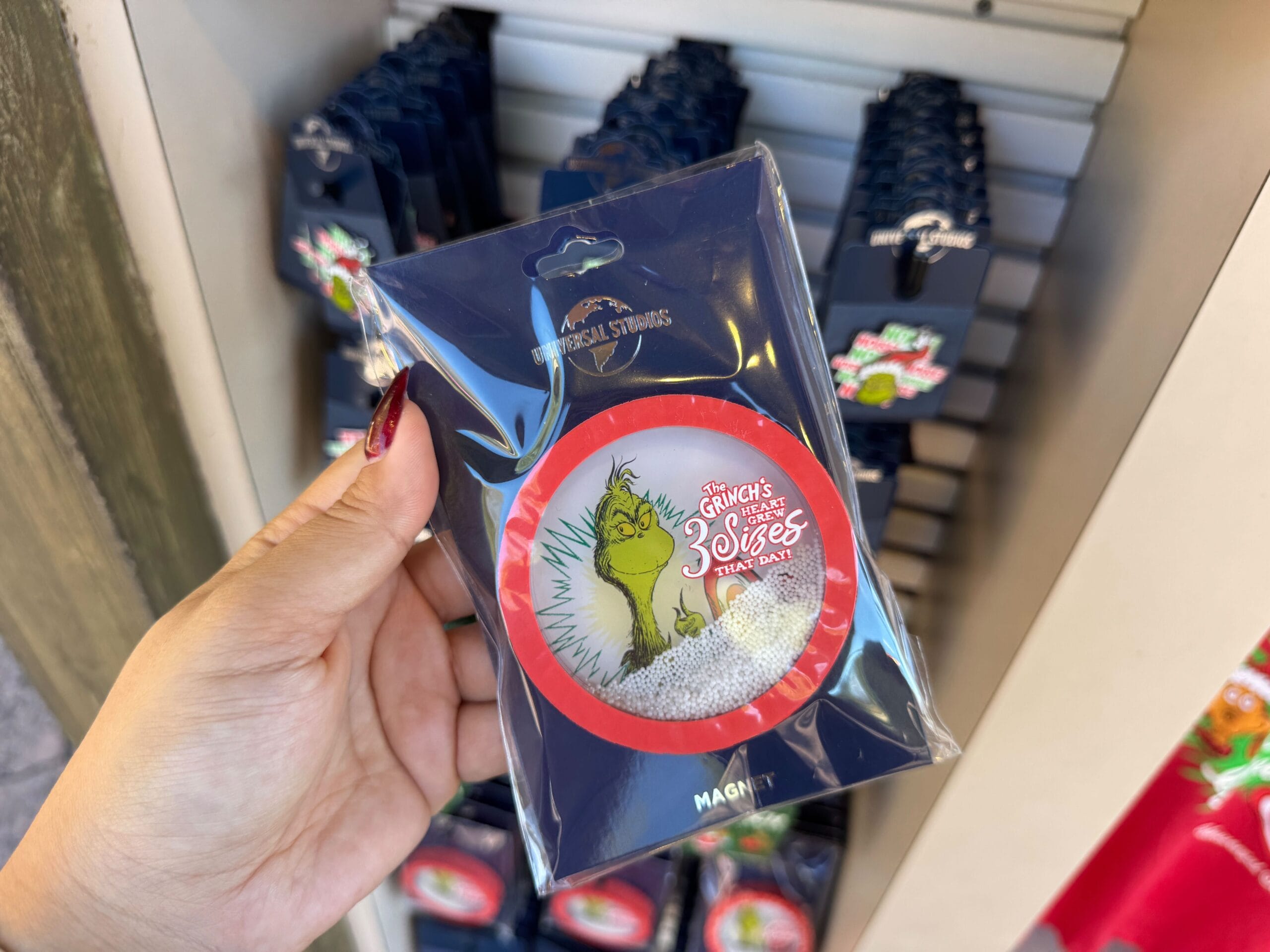A hand holds Grinchmas merch, a magnet reading "Grinchmas at Universal Orlando," in a rack—nothing like Disney castle souvenirs.