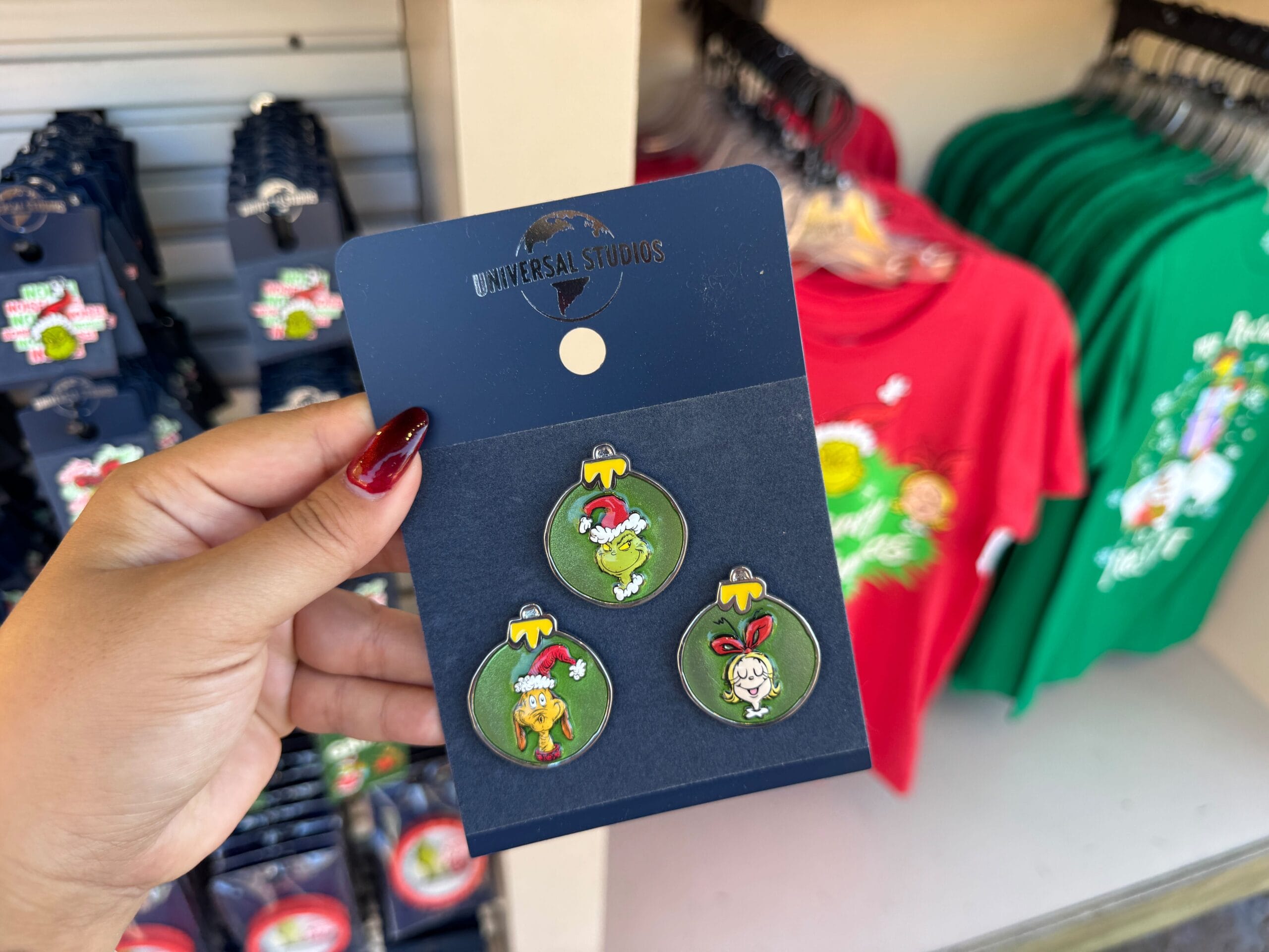 Hand holding Grinchmas enamel pins near festive shirts; though Universal, the scene lacks a Disney castle like Cinderella's at Magic Kingdom.