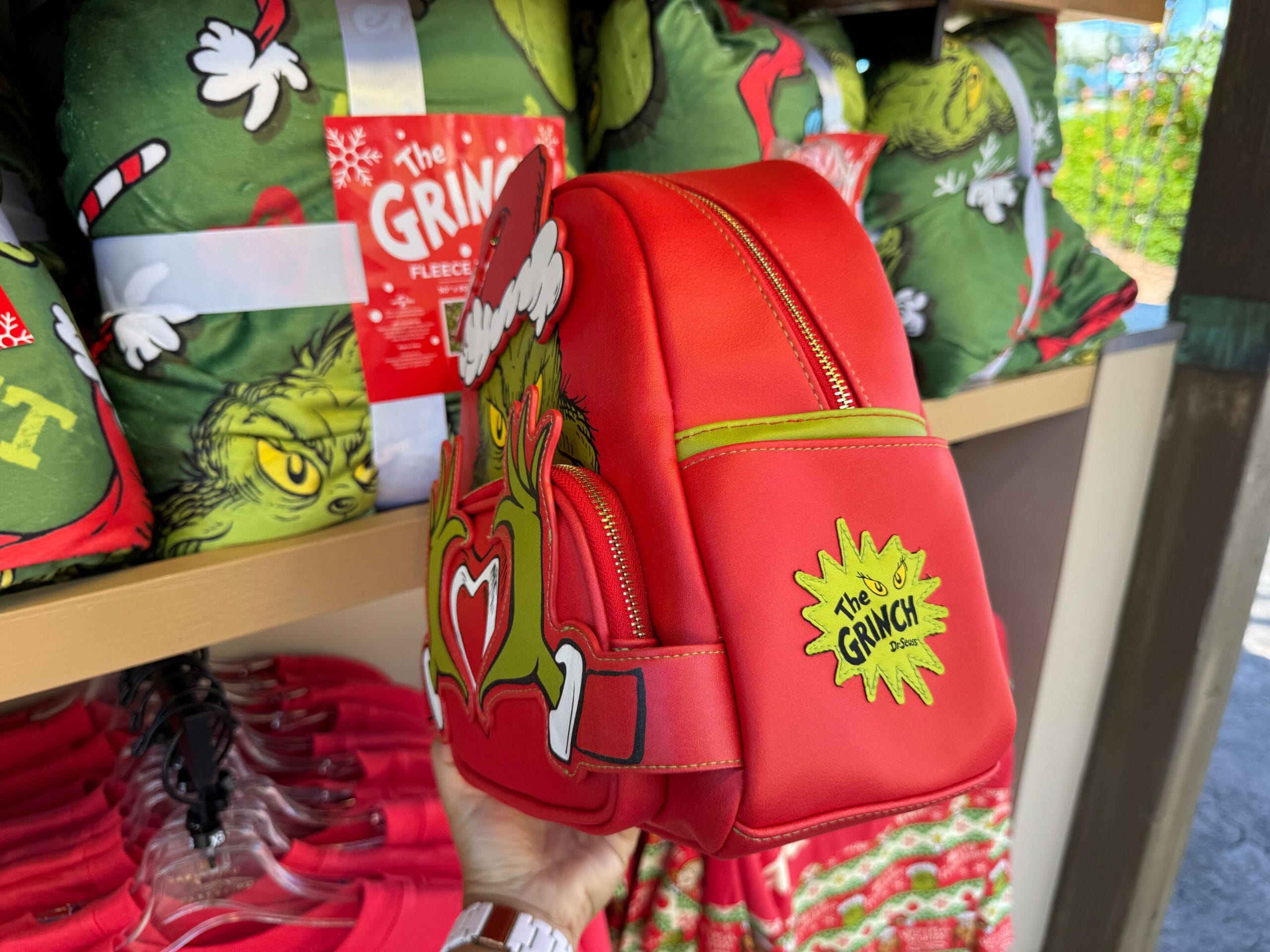 A hand displays a red Grinch-themed backpack at a Universal Orlando store—no Cinderella Castle like at Disney's parks in sight.