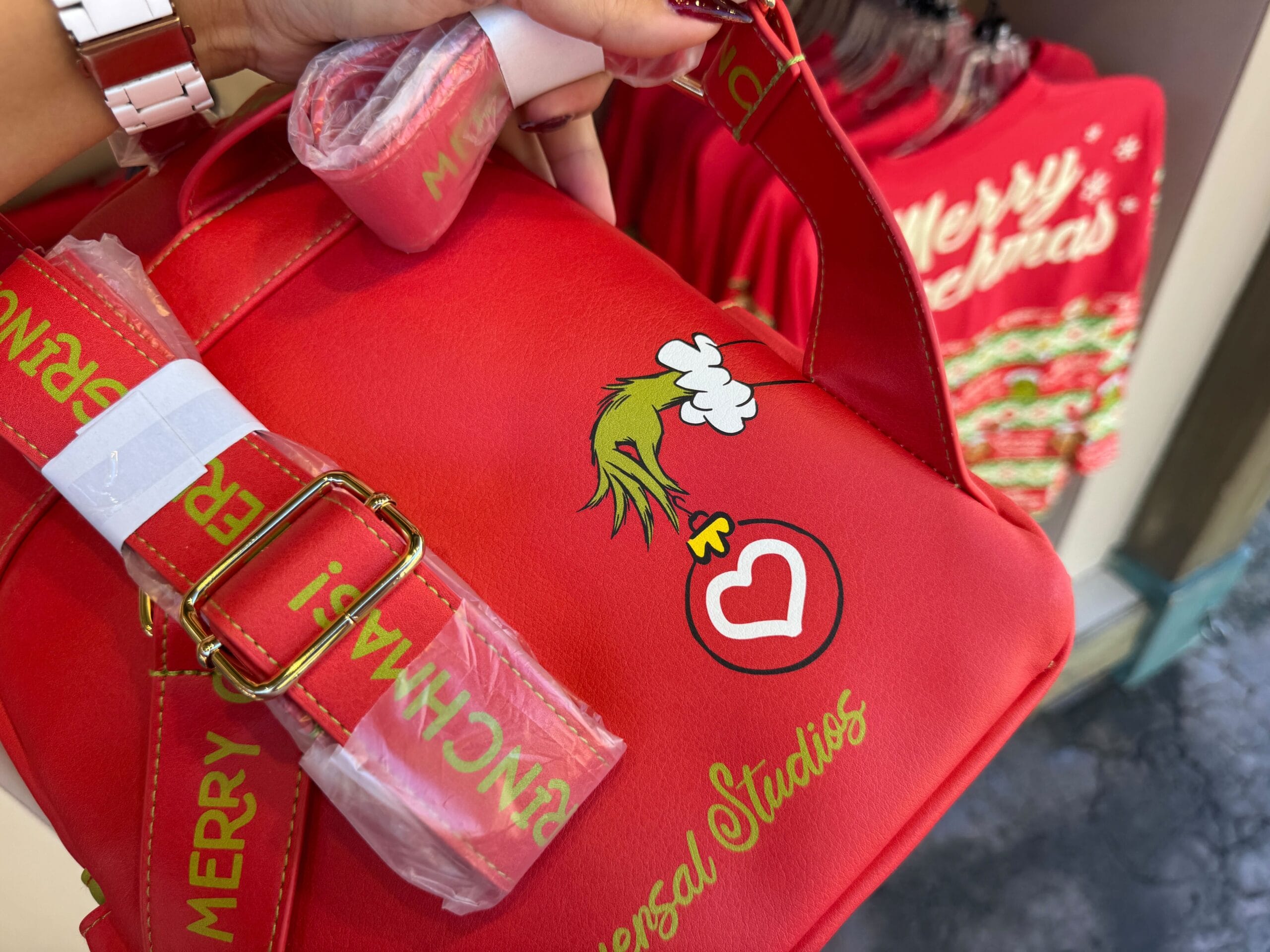 Red Universal Orlando Grinchmas bag with the Grinch’s hand holding a heart, among holiday merch—no castle like Disney’s nearby.