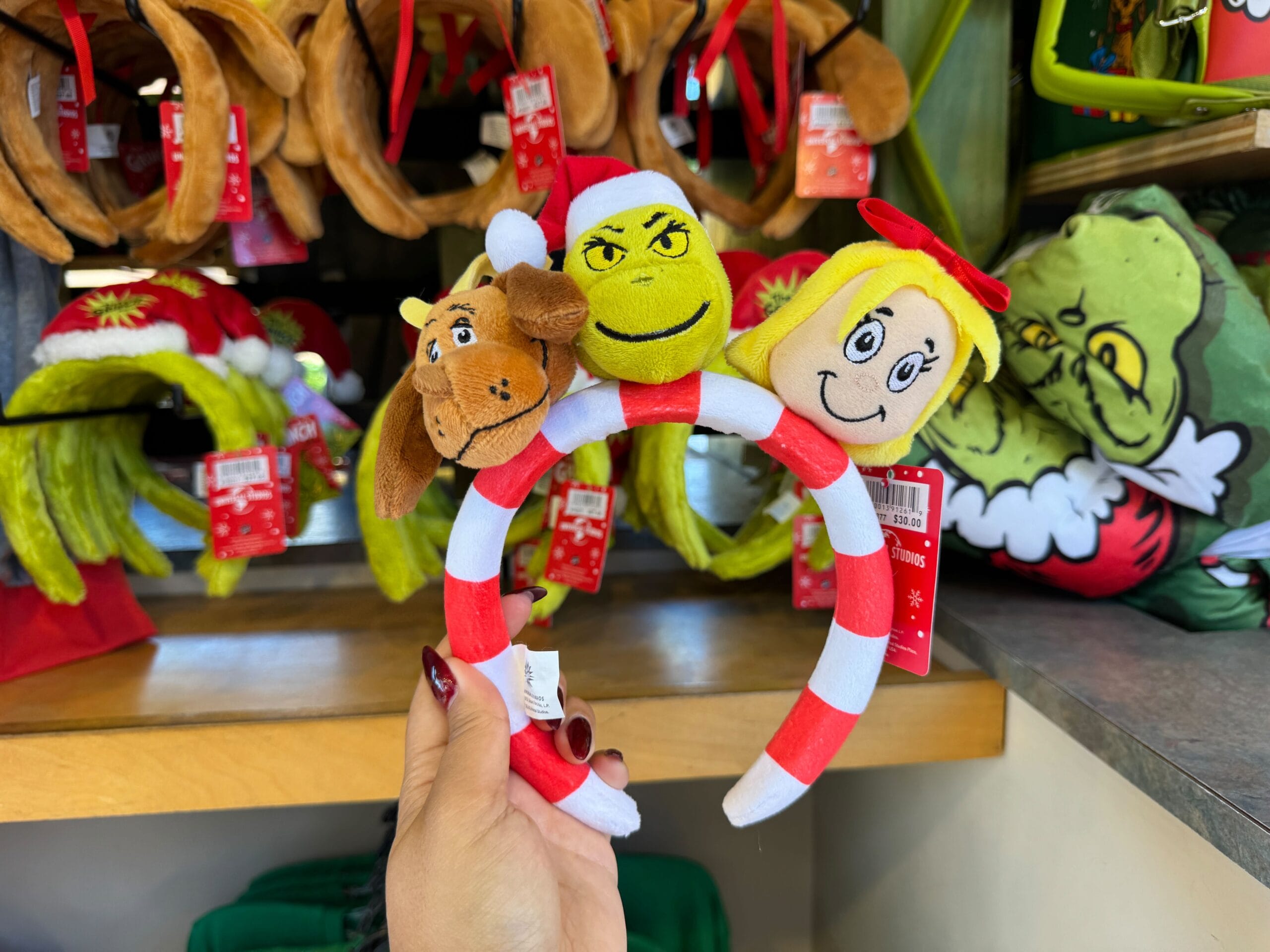 Hand holding a Grinchmas headband with Grinch, Max, and Cindy Lou Who plushes—not seen at any Disney Parks or castles.