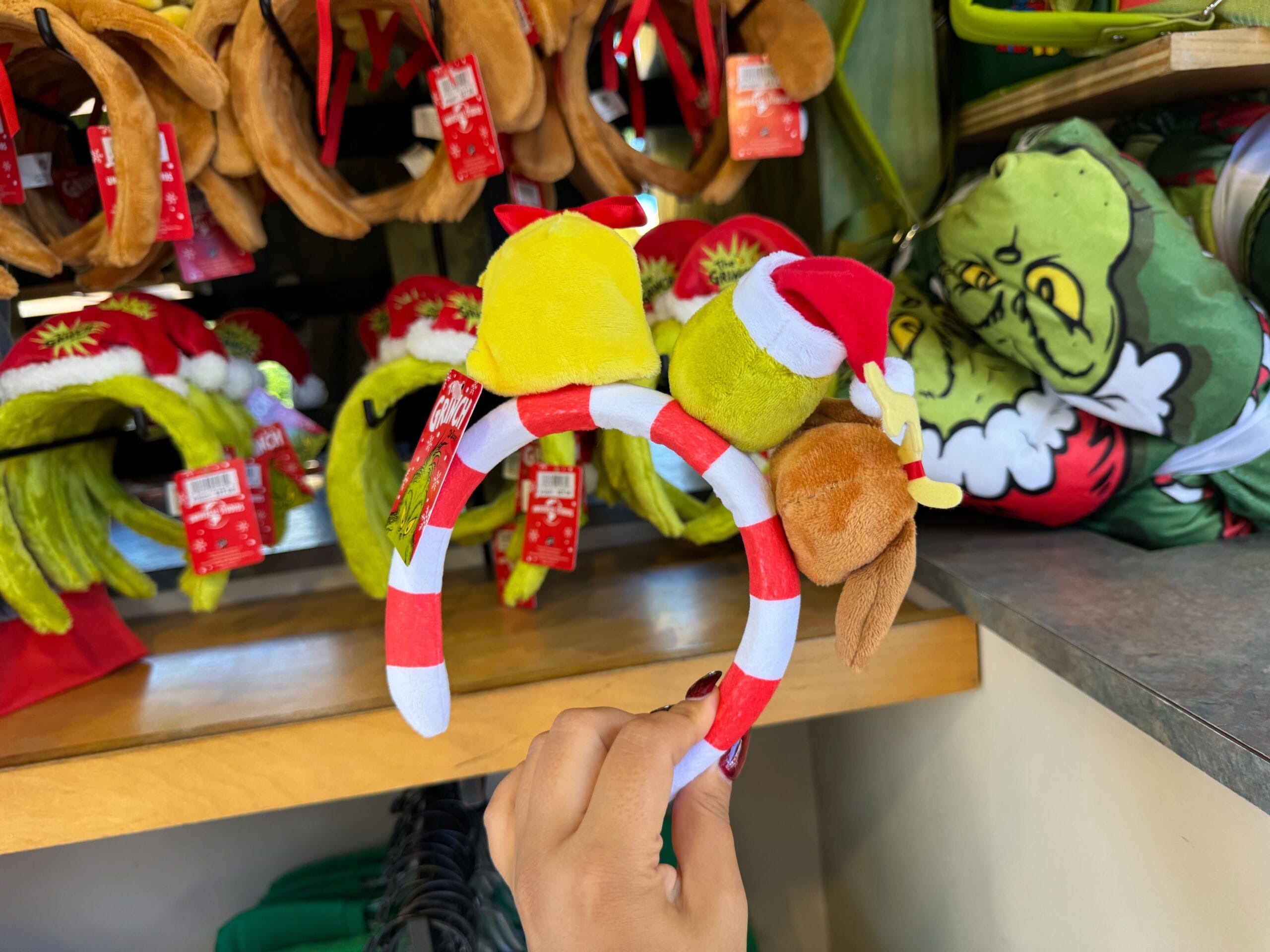 A hand displays Grinchmas merch—a striped headband with plush Grinch, Max, and Santa hat—at Universal Orlando, not a Disney castle.