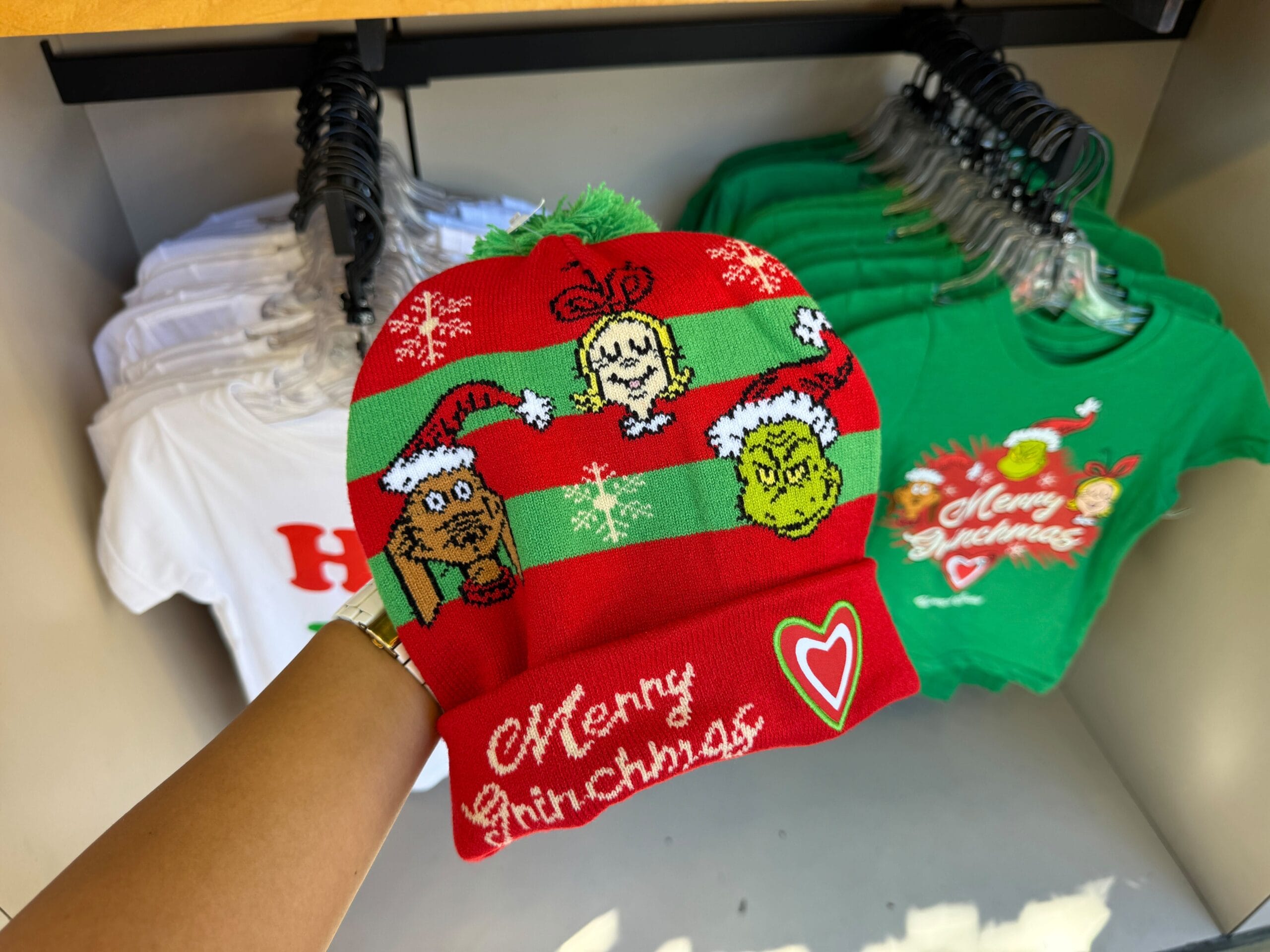 A hand holds a red and green Grinchmas hat with themed merch, displayed before festive shirts—not Cinderella Castle, but Universal.