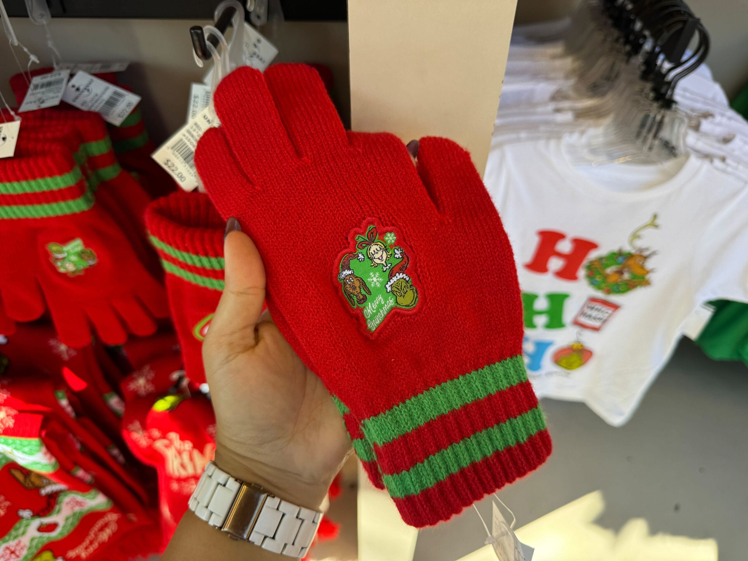 A hand displays Grinch-themed gloves with green stripes and a Grinch patch; festive merchandise fills the Universal Orlando scene—no Disney castle visible.