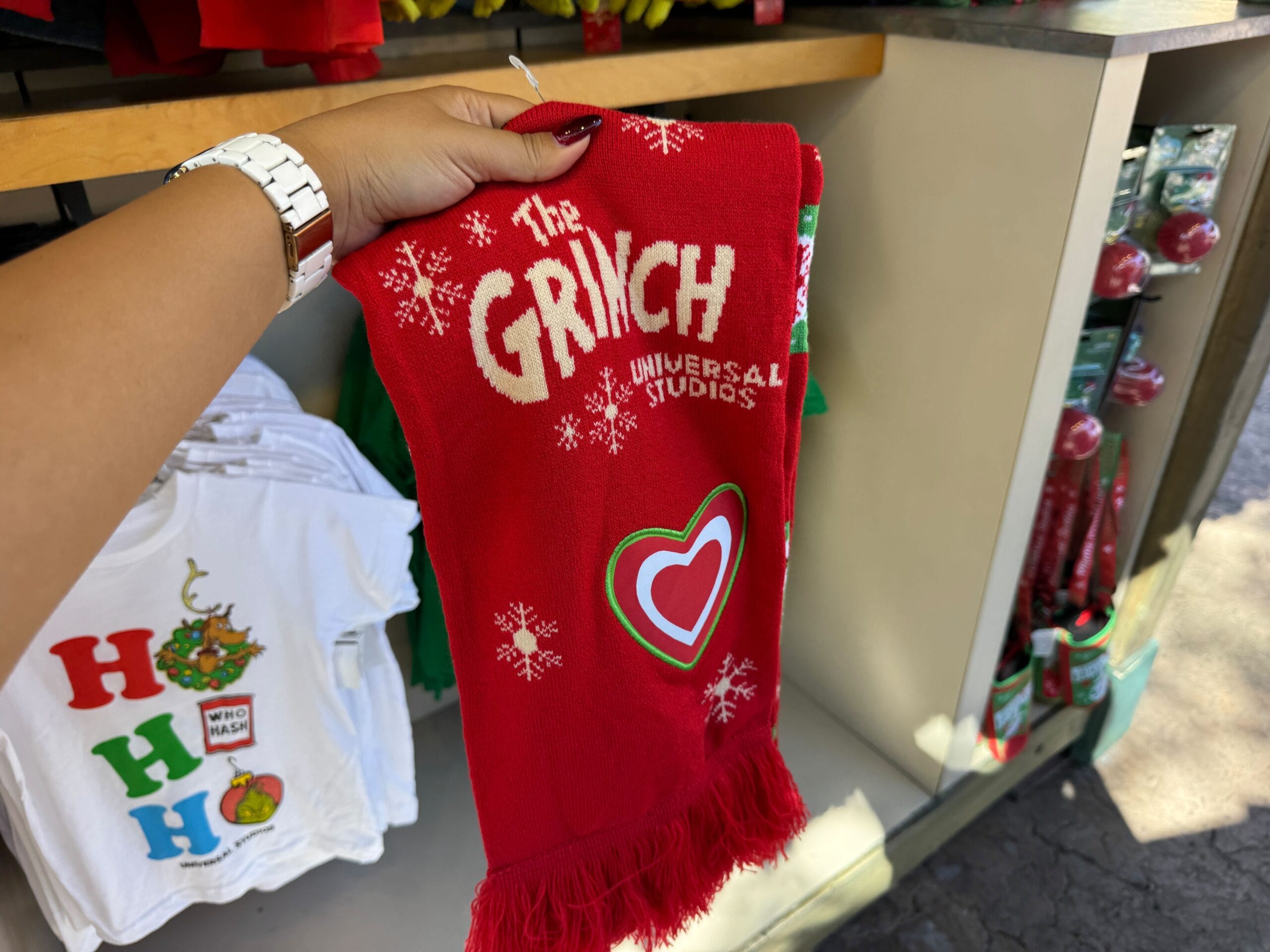 A hand holds Grinchmas merch—a red scarf with "The Grinch Universal Orlando," snowflakes, and a heart, displayed in-store, echoing holiday merch found in Disney Parks but with a Universal twist.