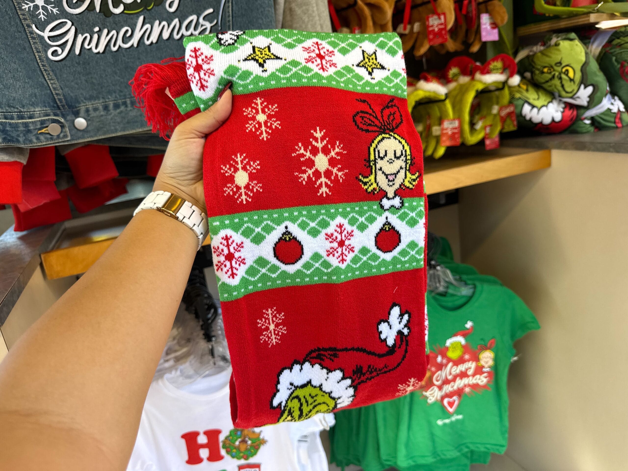 Hand holds a festive Grinchmas scarf featuring The Grinch and Cindy Lou Who with holiday merch, reminiscent of Disney park gifts.