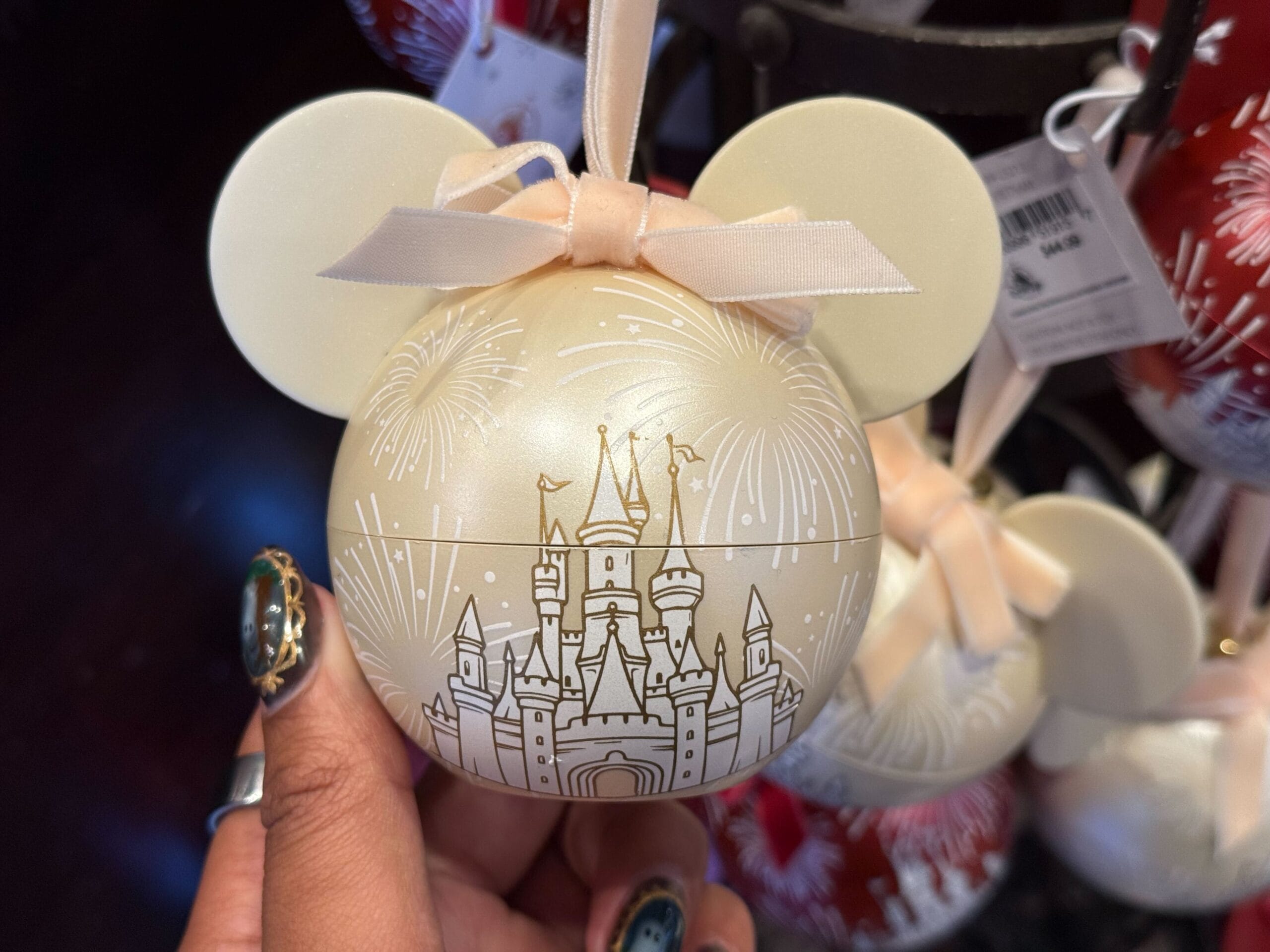 Hand holds cream Mickey ears ornament with pink bow, castle reminiscent of Disneyland’s, fireworks—2025 holiday park exclusive.